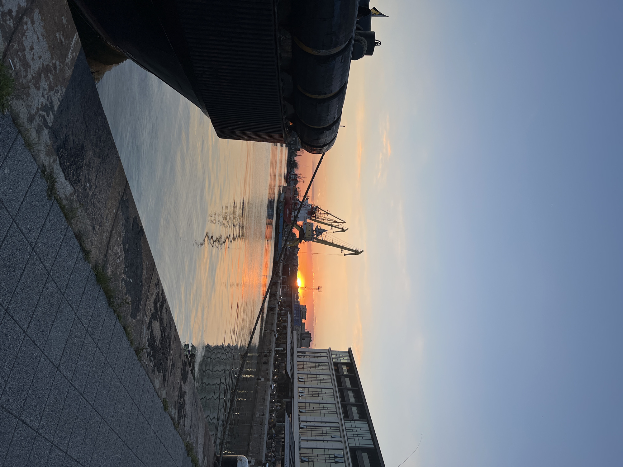 The photograph captures a sunset scene on the waterfront of Burgas, Bulgaria. The sky is a gradient of blues and oranges, reflecting in the calm water. Silhouetted against the sunset are cranes and harbor structures, indicative of a working port. A large ship or dock structure is prominent in the foreground, partially obscuring the view and connected to the shoreline by a mooring rope. On the lower right, a modern building with large windows is visible. The ground is paved with gray tiles, transitioning to a stone wall along the water's edge. There are no people visible.