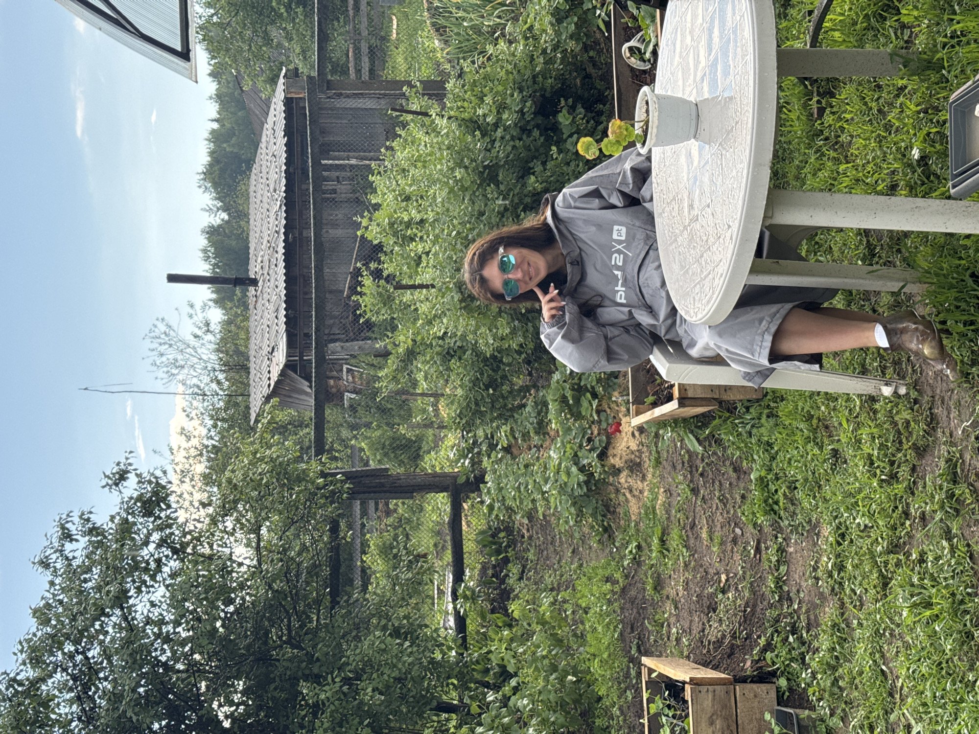 A woman sits at a white round plastic table in a backyard garden. She is wearing a grey hooded garment with a white logo that reads "PH 2XP". She has on sunglasses and is looking towards the camera. Her right hand is resting on her chin. There is a small potted plant on the table. The garden is overgrown with grass and various plants. In the background, there is a wooden fence and a rustic wooden structure with a corrugated metal roof and a chimney pipe. The sky is partly cloudy.