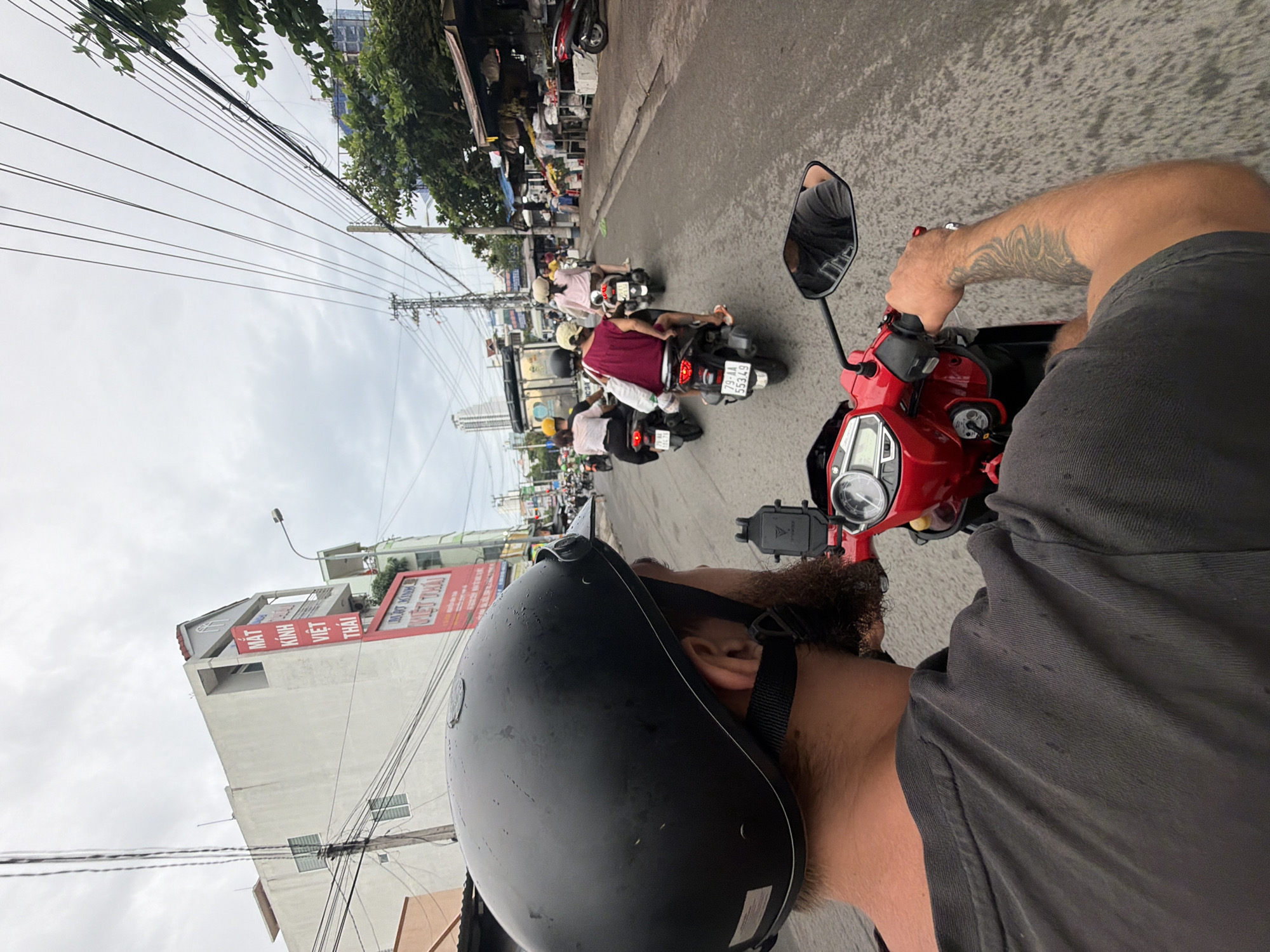 In Nha Trang, Vietnam, a first-person perspective shot captures a person riding a red scooter on a busy street. The rider, wearing a black helmet and a grey t-shirt, has a tattooed arm and a beard. The scooter is equipped with a phone mount. The street is lined with buildings, including one with a large red sign in Vietnamese. Several other scooters, carrying multiple passengers, are also visible on the road. Trees and power lines add to the urban landscape. The sky is overcast, suggesting a cloudy day.