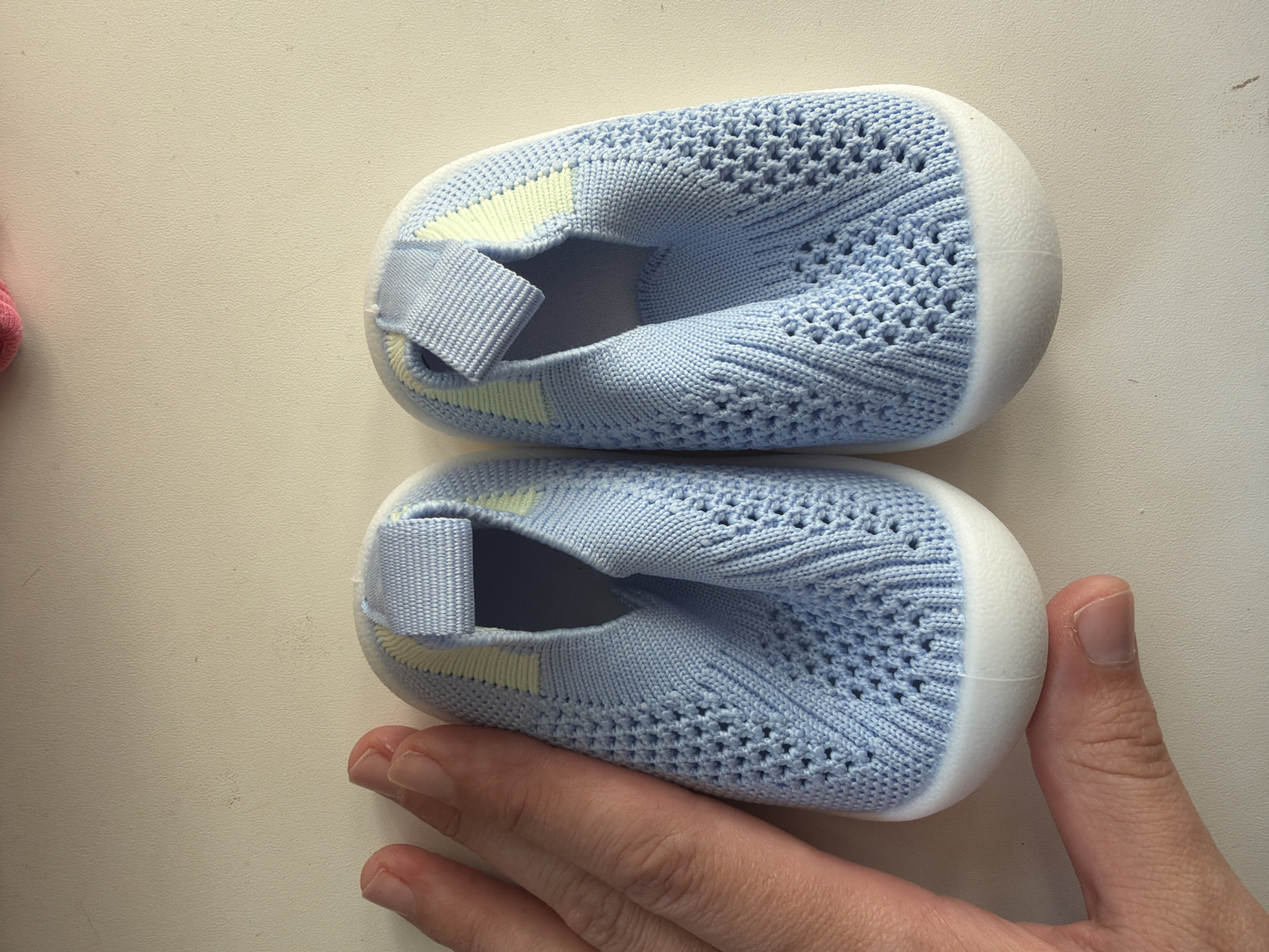 Two light blue knit baby shoes with white soles are positioned on a white surface. A human hand is touching the bottom shoe. The location cannot be determined from the visual information provided.