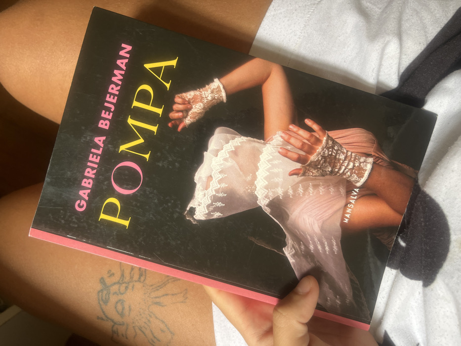 The image displays a close-up, top-down view of a book being held by a person. The book, titled "POMPA" by Gabriela Bejerman, with "MANSALVA" as the publisher, has a dark cover featuring a headless figure in a sheer, lacy, light-colored garment with prominent white fingerless lace gloves. The book is held on its left side by a left hand and rests on the upper thighs of the individual. A line-art tattoo, depicting a stylized creature or figure with multiple appendages, is visible on the upper left thigh. The person is partially clad in white or light-colored fabric, possibly shorts or underwear. No specific location within the city of Buenos Aires, Argentina, can be determined from the visual evidence present in the image.