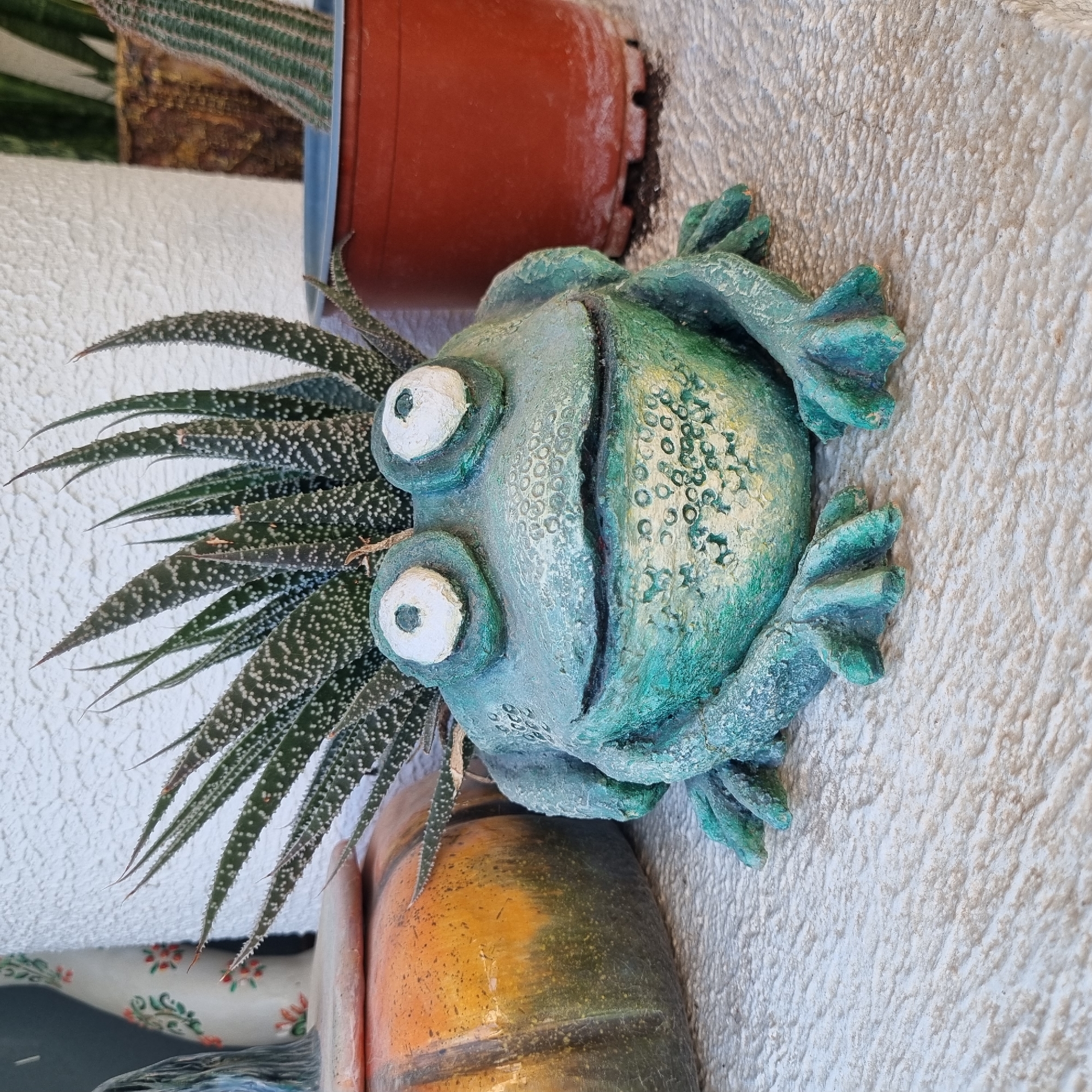 A close-up, eye-level shot shows a frog-shaped planter with a succulent plant growing out of its head. The frog planter is a light green color with yellow accents and large, bulging white eyes with black pupils. The succulent has long, spiky green leaves with white spots. To the left of the frog planter, a portion of a large, orange and green ceramic pumpkin is visible. In the background, to the right of the frog, a brown plastic pot containing a cactus is partially visible. The background consists of a textured white wall. The scene appears to be outdoors, possibly on a patio or balcony in Konyaaltı, Turkey. The focus is sharp on the frog planter and the succulent.