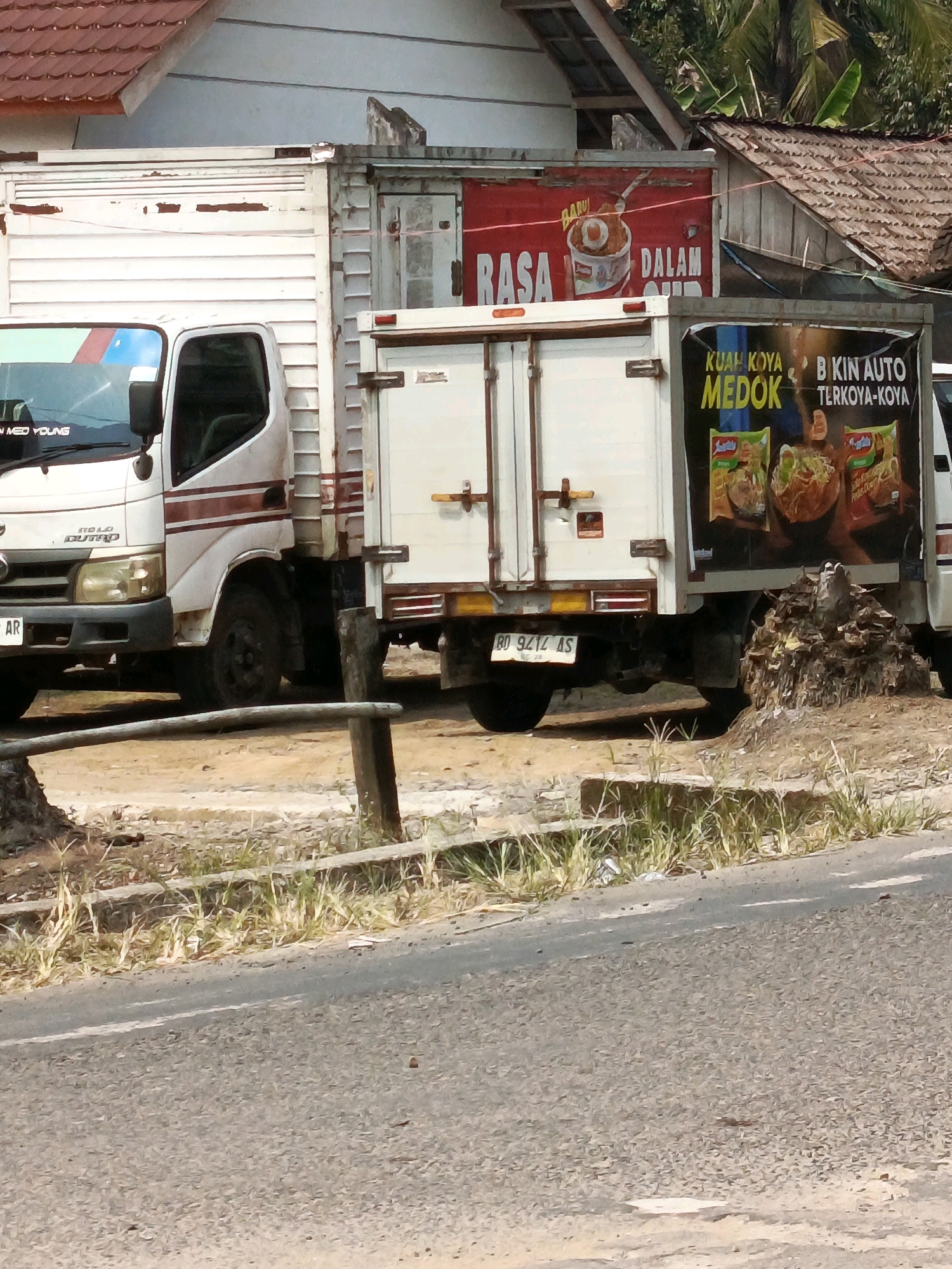 The image depicts a street scene in Wonosari, Indonesia, featuring two trucks parked on a dirt area next to a road. The foreground shows a section of asphalt road with white lane markings and scattered debris. Behind the road, dry grass and some foliage are visible.
The main focus is on two white trucks. The truck on the left is a smaller delivery truck with a visible cab. On its windshield, a white sticker reads "MED YOUNG". The truck on the right is a larger refrigerated truck with a white cargo box. On the side of this cargo box, a large banner displays an advertisement. The banner features images of instant noodle packaging, primarily of the Indomie brand, with text in Indonesian. The text includes "KUAH KOYA MEDOK" and "BI KIN AUTO TERKOYA-KOYA", indicating a promotional message for a type of noodle soup.
Behind the trucks, a building with a red-tiled roof and white walls is partially visible. A red sign with white lettering, featuring an image of a bowl of food, is attached to the side of the building above the trucks. Trees with green foliage are also visible in the background, suggesting a semi-urban or rural setting. The overall atmosphere is sunny, with shadows indicating daylight.