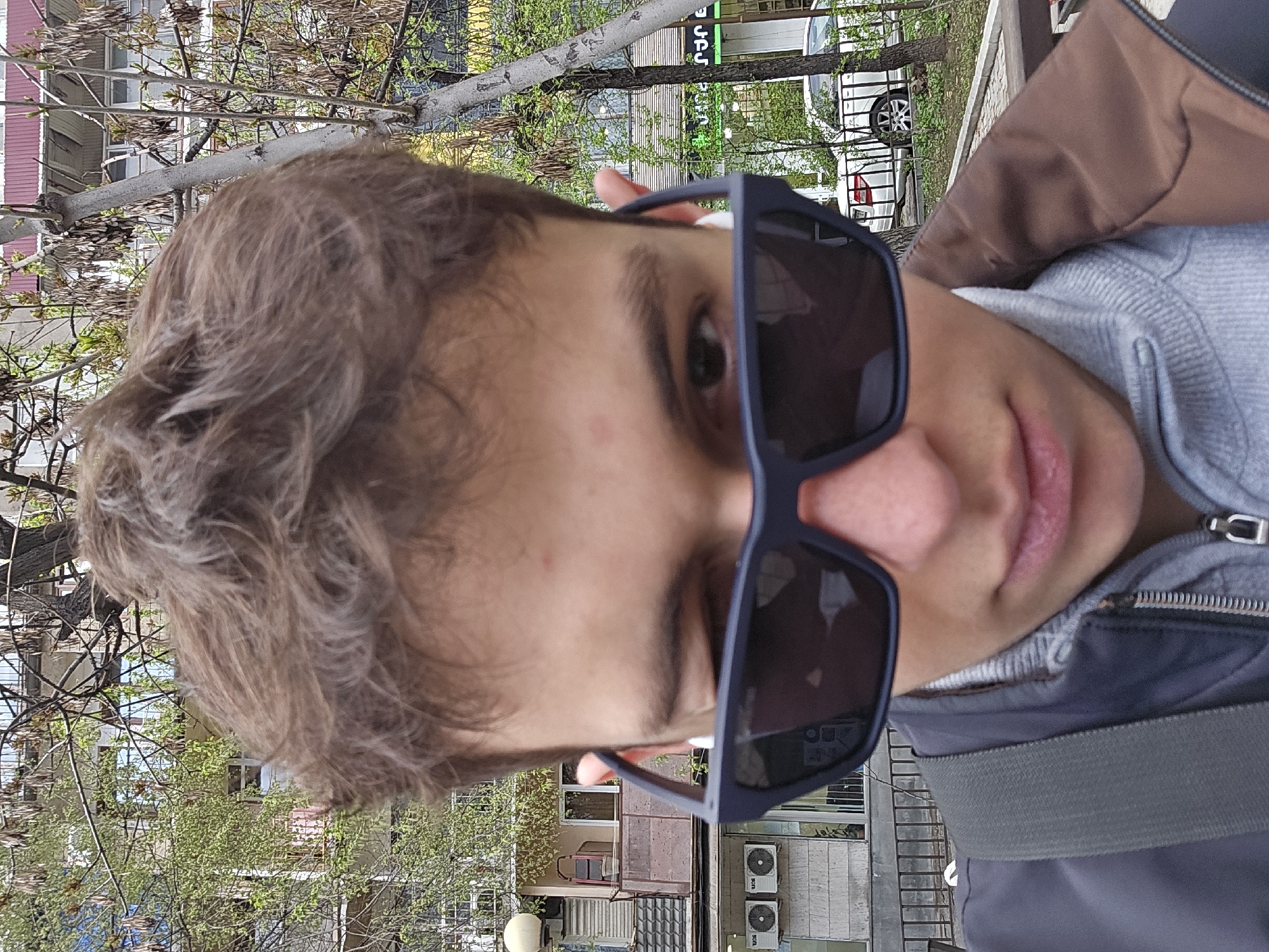 A close-up, slightly rotated shot depicts a young man wearing dark sunglasses. He is dressed in a grey hooded sweatshirt under a dark blue jacket with a brown collar. A grey strap crosses his chest.
Behind him, a city scene is visible. There are trees with fresh green leaves, buildings with windows, and air conditioning units mounted on a wall. A parked white car is also visible.
Based on visual clues, the location is likely Yerevan, Armenia. The architectural style and signage, while partly obscured, align with typical urban elements found in Yerevan.