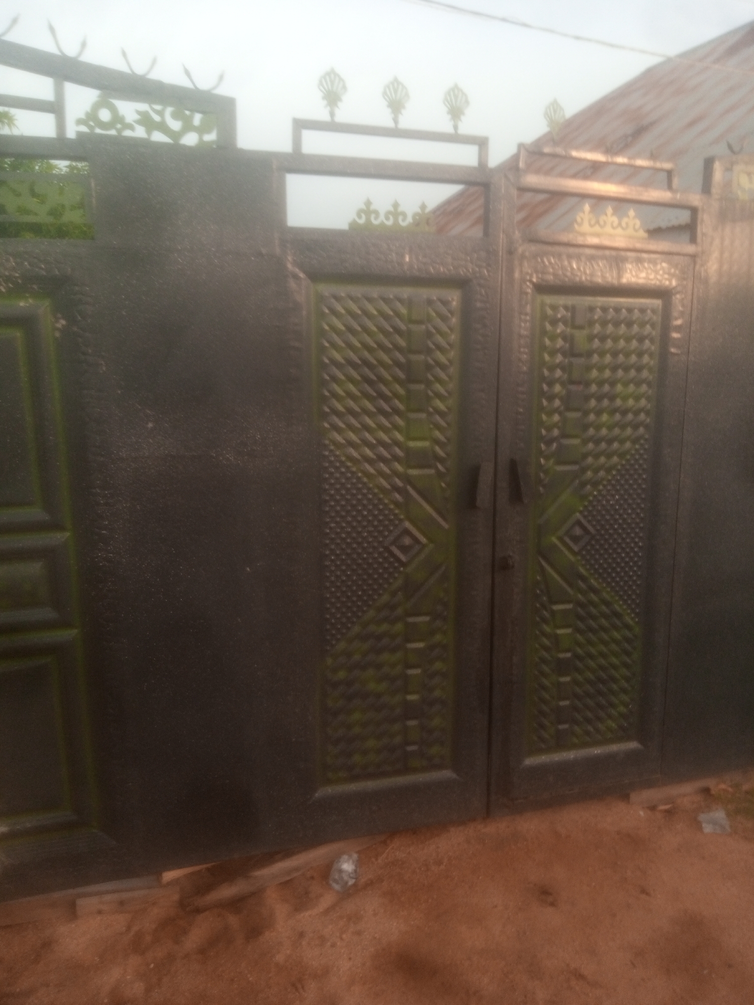 The image shows a closed metal gate in Jalingo, Nigeria. The gate is dark gray with decorative green patterns on the panels. The gate has two sections, each with a handle and a lock. The gate is set in front of a building with a corrugated metal roof. The ground in front of the gate is covered with reddish-brown sand. The sky is overcast.