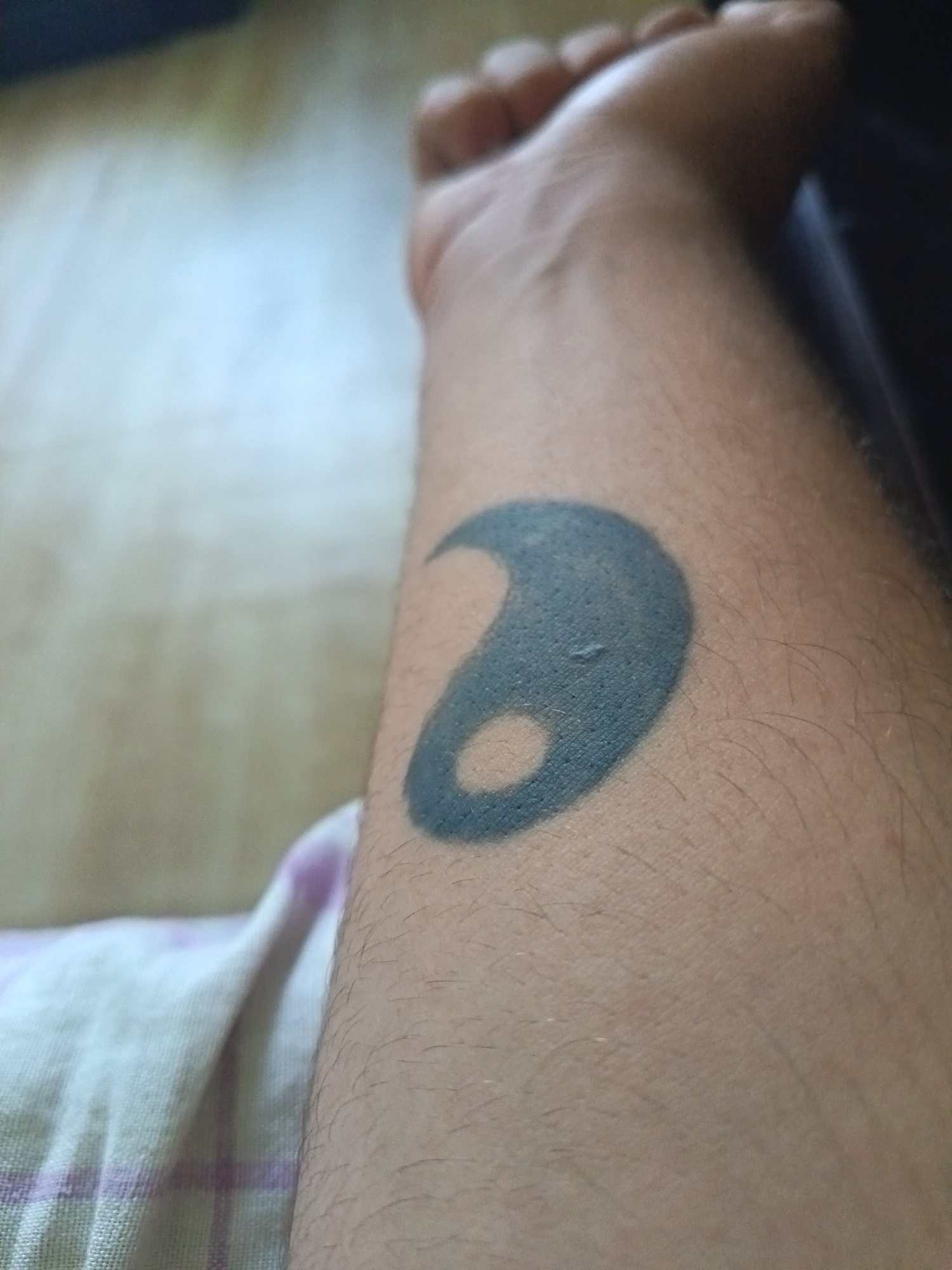A close-up image shows a person's forearm with a black tattoo resembling a stylized crescent shape or part of a Yin and Yang symbol. The skin is light-colored with fine hairs. The arm rests against a patterned fabric with white and pink squares. The background is blurry and indistinct, showing a light-colored wooden surface. The location in Drenovets, Bulgaria is undetermined as there are no local indicators.