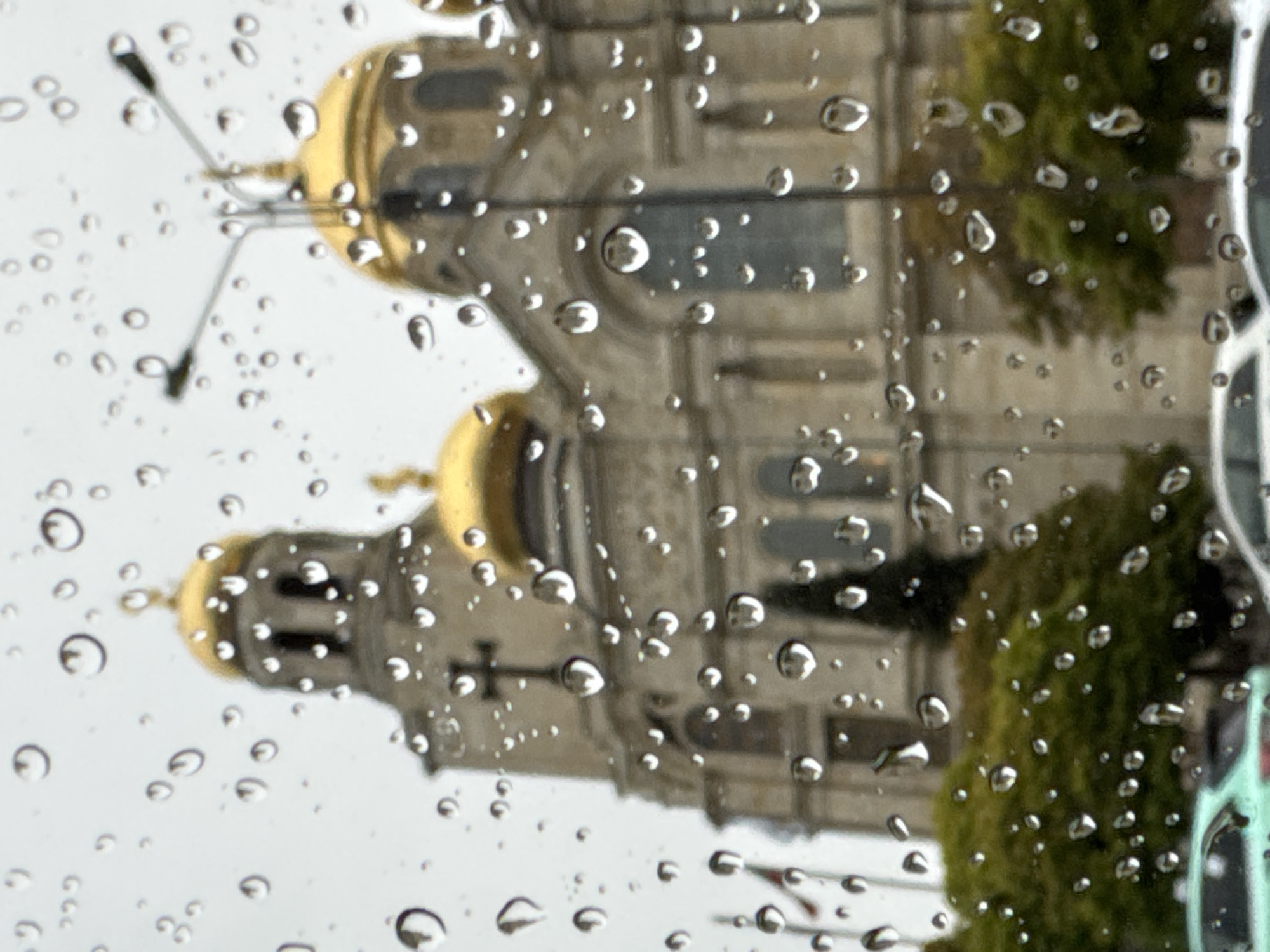 An image captures a view of a grand architectural structure, likely a cathedral or church, seen through a rain-streaked window. The image is rotated 90 degrees clockwise, presenting the building on its side.
The main subject is a large stone building, featuring multiple golden-domed towers characteristic of Orthodox Christian architecture. One prominent dome and a spire with a cross are clearly visible on the left side of the rotated image. The building's facade appears to be made of light-colored stone, with intricate details and windows, though these are blurred by the water droplets.
In the foreground, numerous clear water droplets of varying sizes cling to a transparent surface, distorting and blurring the background scene. This creates an impressionistic effect, with the building's features softened and diffused. Hints of green foliage from trees or bushes are visible to the right of the main building, also blurred by the rain. A street light pole can be discerned extending from the upper left of the rotated image.
The weather is distinctly rainy or has recently rained, as indicated by the water droplets. The sky appears overcast, suggesting daytime with diffused light. The architectural style, particularly the golden domes and cross, strongly points to an Orthodox church. Given the location context of Varna, Bulgaria, this structure is likely The Dormition of the Theotokos Cathedral. No people or visible text are present in the image. The unique perspective through the wet glass offers an abstract and atmospheric view of the urban scene.