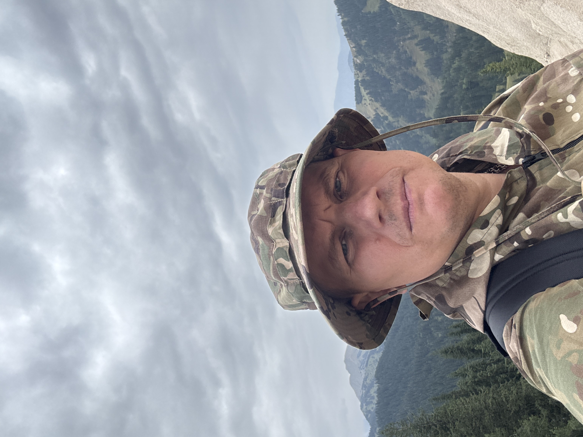 A man wearing a camouflage hat and jacket takes a selfie outdoors, with a backdrop of a cloudy sky and mountains covered in green trees. He is wearing earbuds. The man appears to be on a hike or outdoors excursion. The rocky texture of a light-colored rock formation is visible on the right side of the image. The scene is likely a mountainous region near Almaty, Kazakhstan, given the vegetation and mountainous terrain.
