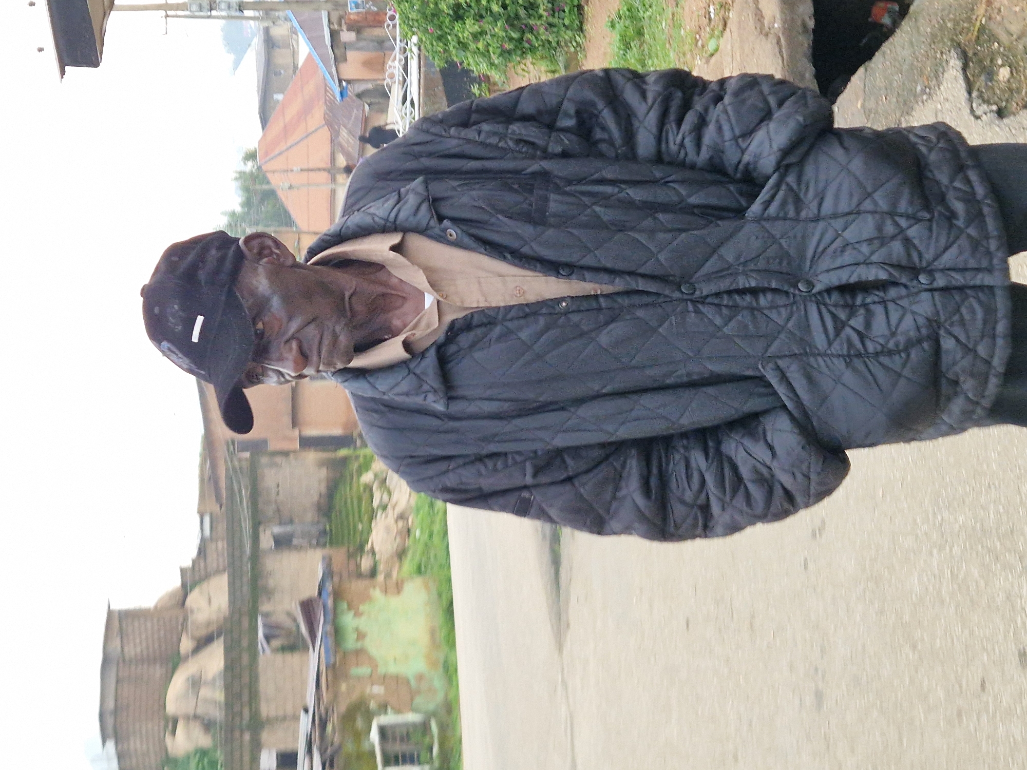 An elderly African man wearing a black baseball cap and a quilted black jacket over a tan shirt stands on a paved road. The man's face is visible, and his hands are in the pockets of his jacket. In the background, there are buildings with corrugated metal roofs, some of which appear weathered. To the left, there are structures made of concrete blocks and some vegetation. To the right, there is a ditch or drain along the side of the road with some greenery. The sky is overcast. The scene depicts a street in Kabong, Nigeria.
