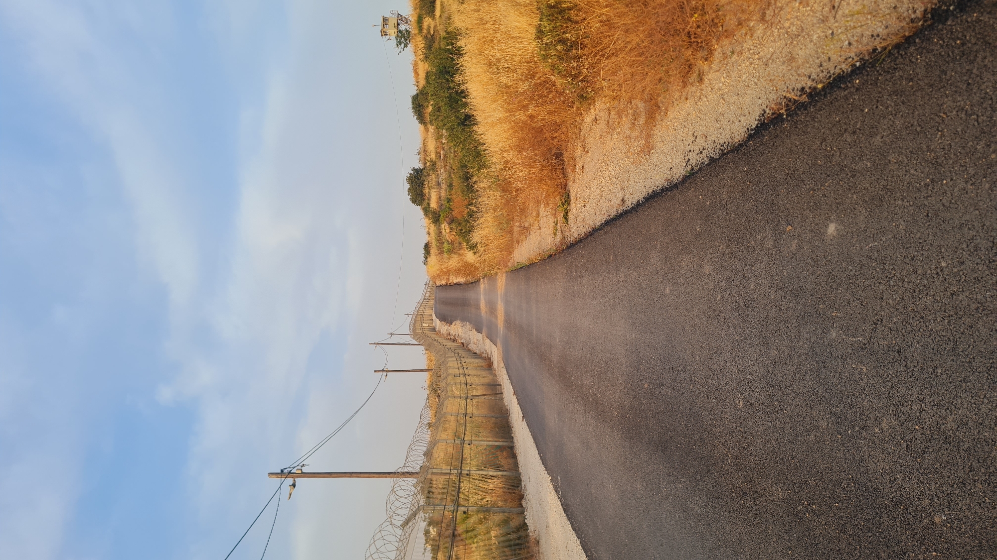 The image shows a narrow asphalt road cutting through a landscape. A fence runs along the left side of the road, reinforced with barbed wire. Wooden utility poles stand alongside the fence, with power lines visible against a blue sky. On the right side of the road, dry, golden vegetation covers the slope. In the distance, a watchtower stands on the hilltop. The road appears to stretch into the distance. The location is in the city of Lachish Regional Council, Israel.