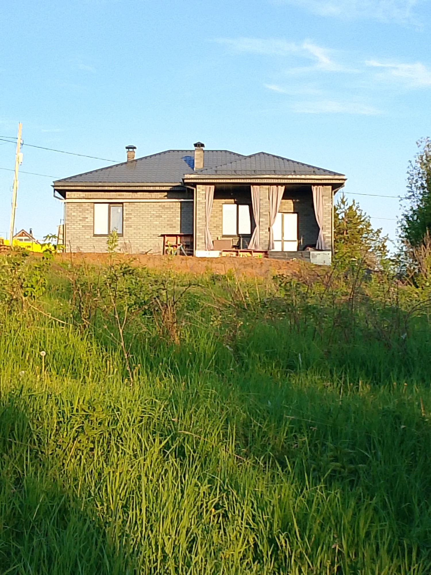 A single-story brick house with a dark gray roof is situated on a small rise. The house has two visible chimneys, one on the left and one on the right. Gray curtains are hanging on the right side of the house. A wooden table and chairs are visible on the patio. Tall green grass and bushes cover the foreground. A utility pole is visible in the background on the left. The sky is blue with few clouds. The location is Прикамский, Russia.