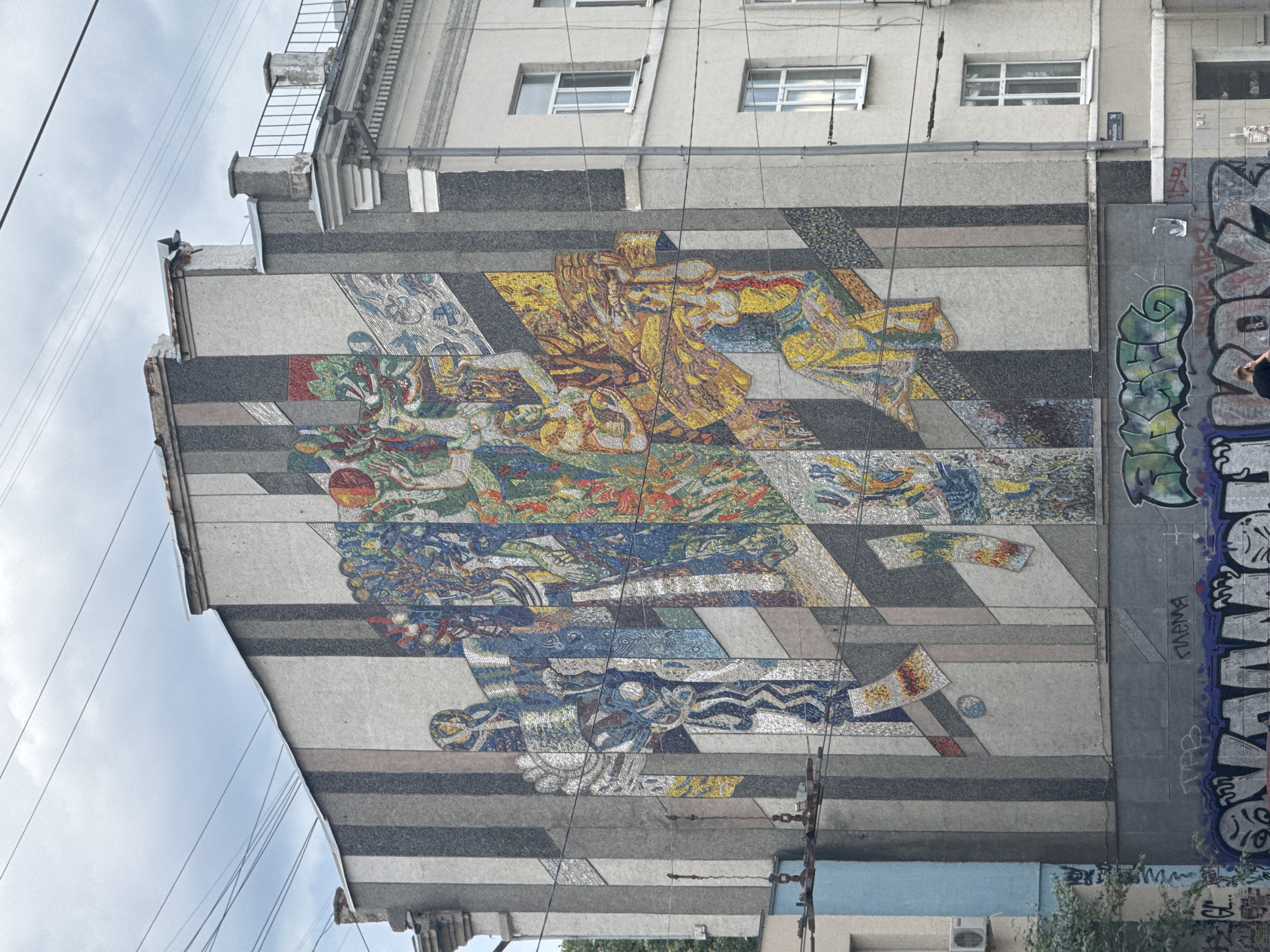 The image shows a large, colorful mosaic mural covering the side of a building in Yekaterinburg, Russia. The mural depicts abstract figures and geometric patterns in vibrant blues, yellows, reds, and greens, set against a background of vertical gray and beige stripes. The mosaic is comprised of small, tessellated tiles.
The building itself is multi-story and appears to be residential or mixed-use, with windows visible on the right side of the frame. Utility wires crisscross in front of the mural and the building. Below the mural, the lower part of the building is covered in graffiti.
To the left of the mosaic, a portion of a lighter colored building with a blue section is visible, along with some greenery and an air conditioning unit. The sky above is overcast.
A sign on the right side of the building, below the windows, reads "МАЛЫШЕВА 76" (Malysheva 76), indicating the address.