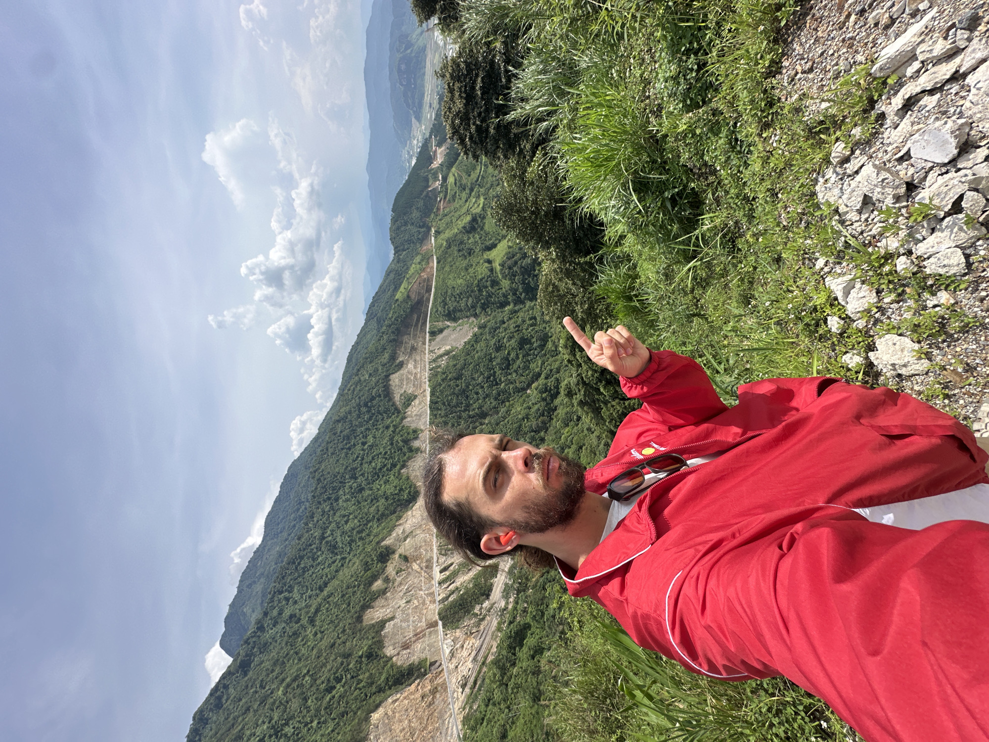 The image captures a scenic mountainous landscape, likely in Muong Than, Vietnam. A man, appearing to be in his 20s or 30s, is prominently featured in the foreground, facing towards the distant view. He has a beard and dark hair, and is wearing a bright red jacket over a white t-shirt. Orange earplugs are visible in his ears, and sunglasses rest on the collar of his jacket. With his right hand, he is pointing his index finger upwards and slightly to the left, directing attention towards the expansive scenery. His expression is neutral and contemplative.
He stands on a rugged mountain slope, characterized by patches of green grass and loose rocks and gravel underfoot. The background unfolds into a vast panorama of rolling mountains, densely covered in lush green trees and vegetation. A prominent feature of the landscape is a wide, winding road cut into the mountainside, showcasing significant terraced construction. In the far distance, multiple layers of mountains fade into a hazy, light blue, suggesting great depth and scale. The sky above is partly cloudy, with scattered white clouds against a clear, light blue expanse. The bright lighting indicates it is daytime, likely late morning or early afternoon, with sunny weather. The scene conveys the grandeur of nature combined with human infrastructure, suggesting exploration or travel through a magnificent, rugged region. There is no visible text in the image.