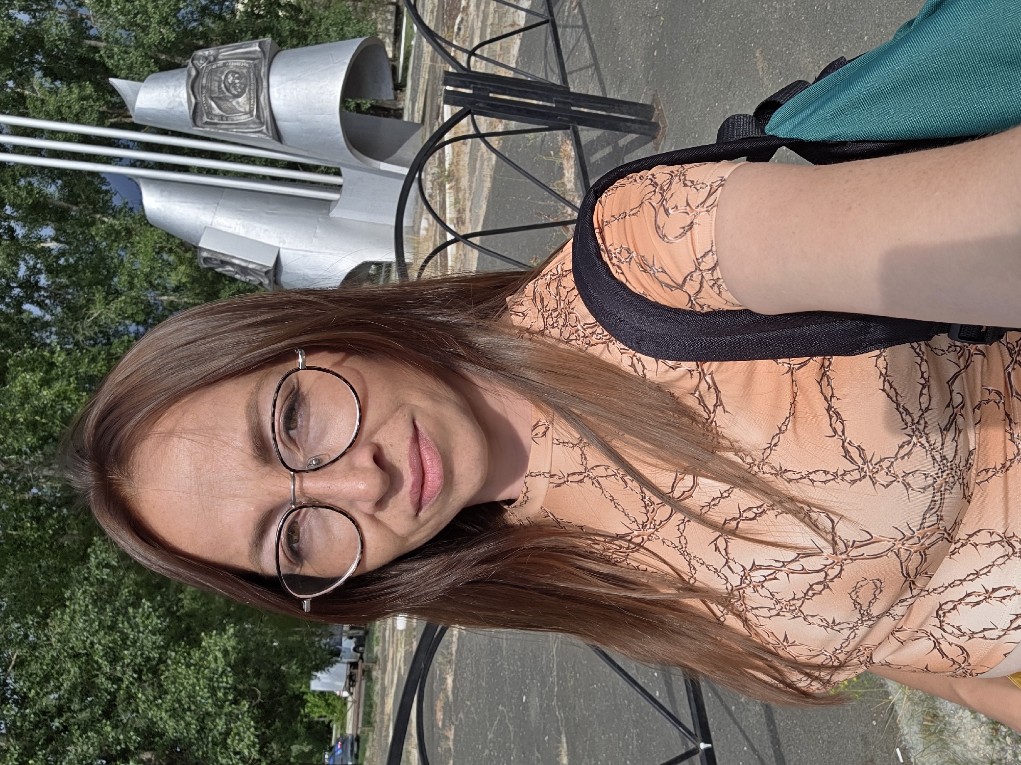 A woman with long, brown hair and glasses is captured in a selfie outdoors. She is wearing an orange top with a dark brown, thorny vine pattern and a black backpack with a teal bag attached. Behind her, a black metal fence separates her from a grey, abstract sculpture and lush green trees. The ground appears to be paved. The scene suggests a sunny day in an urban park or public space.