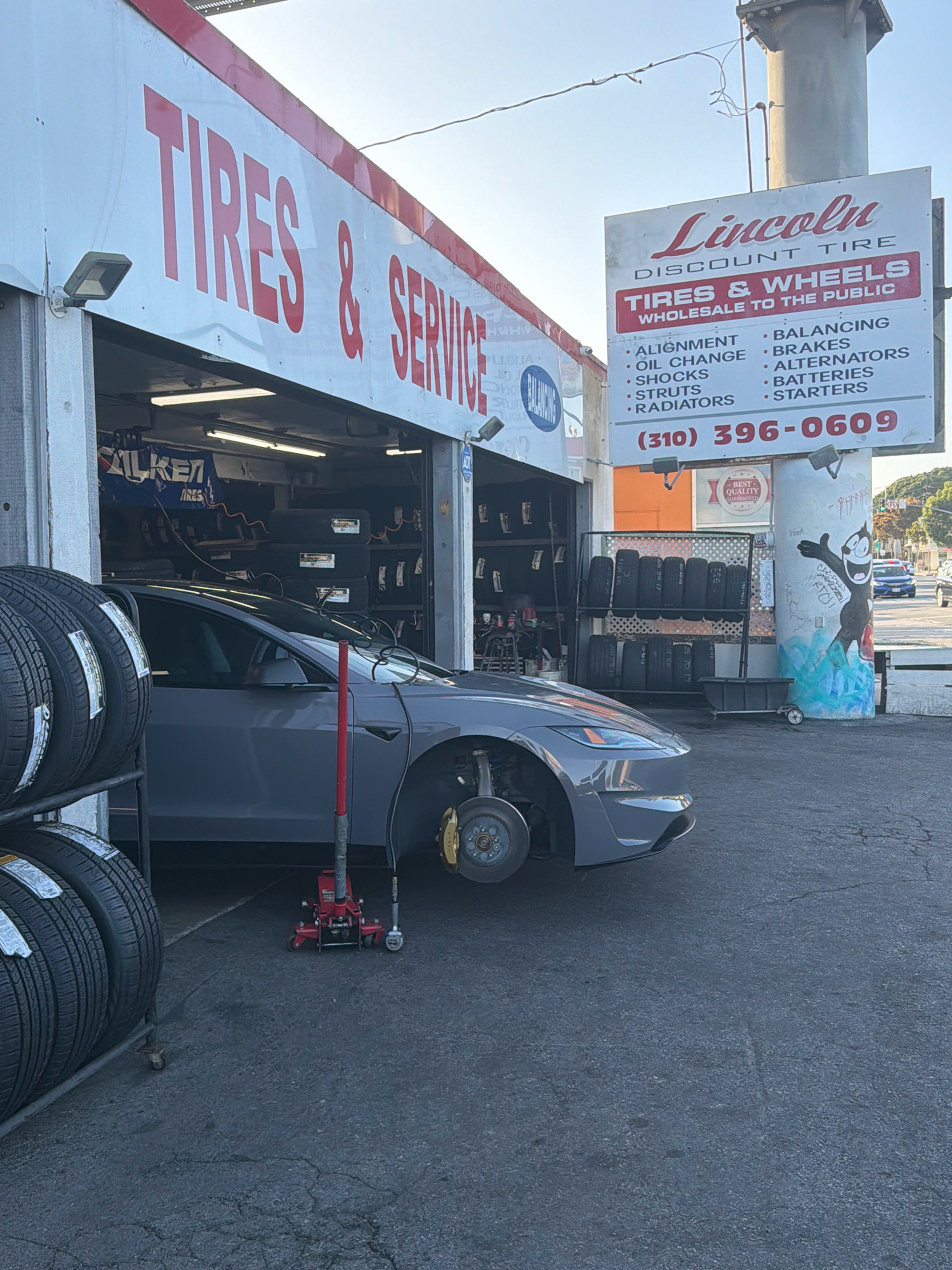 A gray Tesla Model 3 is parked in front of a tire shop with its front right wheel removed. The car is positioned to the right of a stack of new tires and a red hydraulic jack is placed under the vehicle, suggesting it is either being worked on or has recently been worked on. The tire shop has a red and white sign that reads "TIRES & SERVICE" and another sign for "Lincoln DISCOUNT TIRE" listing services and a phone number. Inside the open bays of the shop, more tires are visible, along with a blue banner that says "FALKEN TIRES". The environment appears to be an urban setting, possibly in the city of Los Angeles, given the style of the buildings and the presence of a Los Angeles Police Department vehicle in the distance on the street. The sky is clear and bright, indicating daylight. There are no people visible in the immediate vicinity of the car or the tire shop.