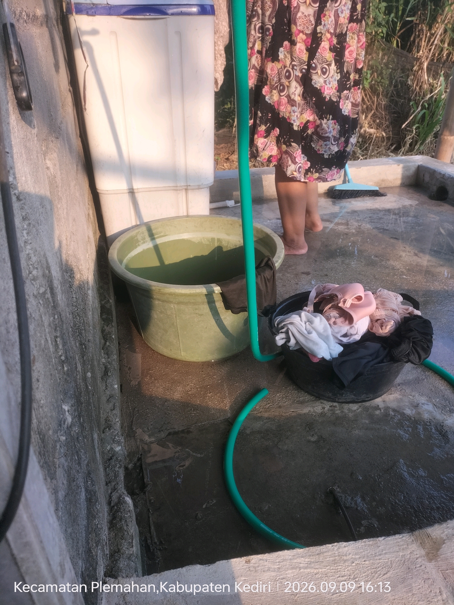In the city of Plemahan, Indonesia, at 4:13 PM on September 9, 2026, a close-up shot captures an outdoor laundry area. A white washing machine is partially visible to the left, with a green hose running from it towards a large, olive-green basin filled with water. Beside the basin, a black bucket is overflowing with laundry, including pink bras and various colored garments. A person, wearing a black skirt with a floral print featuring owls and flowers, stands with their legs visible, suggesting they are engaged in the laundry process. The ground is wet and reflects the sunlight, indicating recent activity. The scene is bordered by concrete structures and overgrown vegetation, bathed in the warm light of the late afternoon sun.
