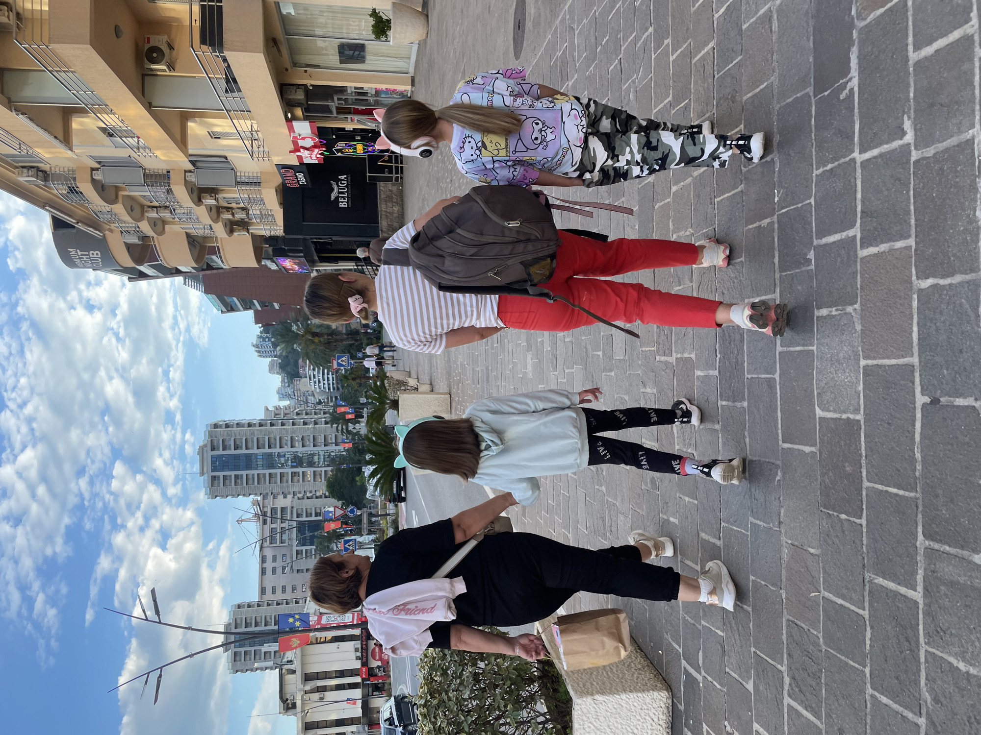 The image displays an urban street scene in Budva, Montenegro, captured on a bright, partially cloudy day. Four females are walking on a gray paved sidewalk composed of rectangular blocks.
On the far left, a woman with light brown hair wears a black top and pants, carrying a light-colored crossbody bag and a brown paper bag. A white sweatshirt with the word "Friend" is draped over her shoulder. To her right, a young girl with long brown hair wears a light blue hoodie with cat ears, black leggings prominently featuring the word "LIVE" on the sides, and white sneakers.
Further right, a woman with light brown hair, styled with a pink hair clip, wears a striped t-shirt (light brown and white) and red pants. She carries a dark backpack and sneakers. The rightmost individual is a young girl with long blonde hair, dressed in a colorful t-shirt with cartoon characters, including visible text like "innamoroll" and "mor." She wears camouflage pants and white sneakers, accessorized with pink cat-ear headphones.
The background features multi-story buildings with balconies and air conditioning units. Several shopfronts are visible, including signs for "GRAND CASINO" and "BELUGA," and a sign indicating a "PREMIUM NIGHT CLUB." In the distance, on the left side of the street, more buildings, palm trees, and streetlights are visible against a bright blue sky dotted with white clouds. Another sign partially reads "tel Imate prijatelje!". The individuals appear to be casually dressed, strolling through what seems to be a commercial or tourist district.