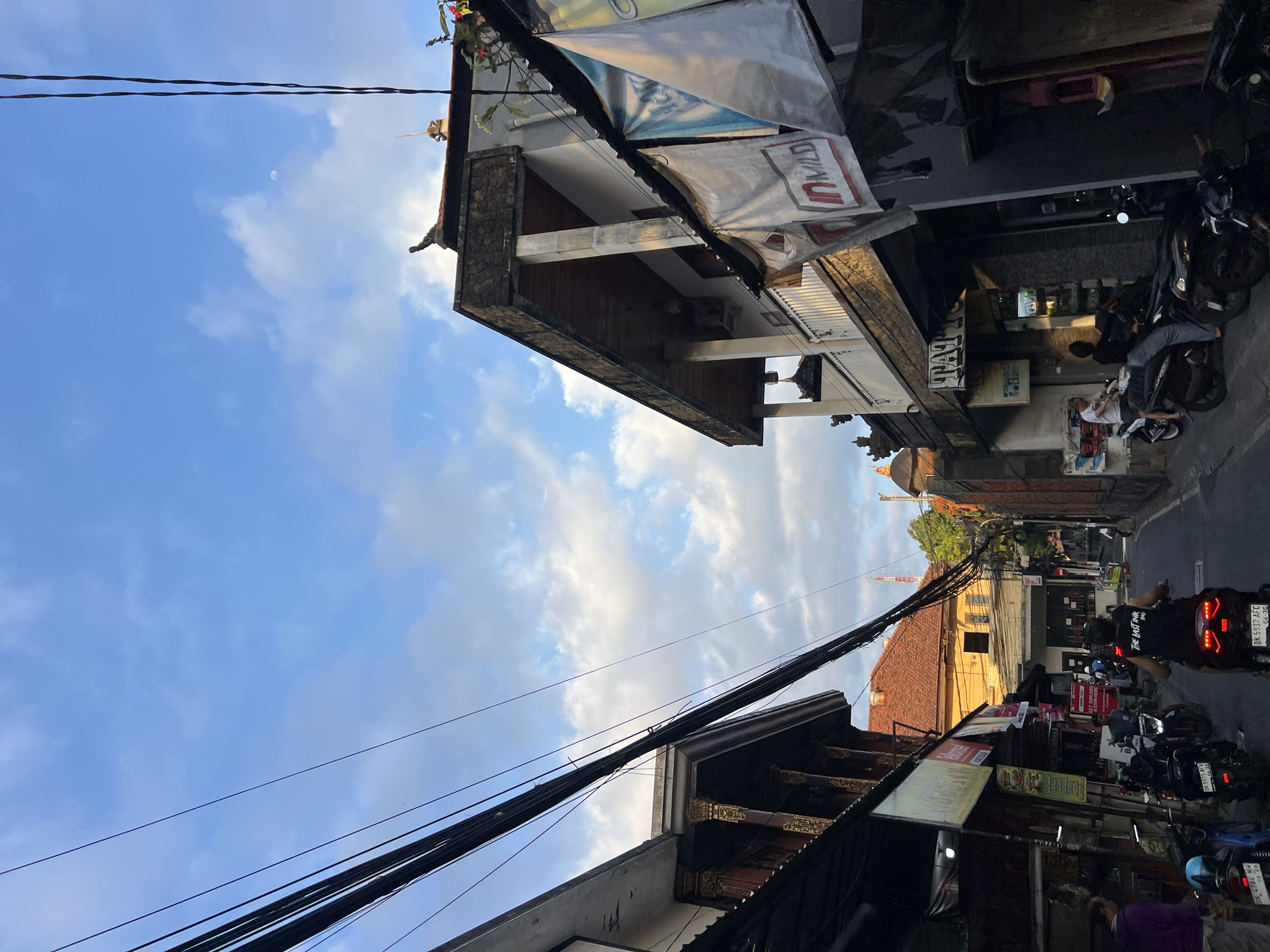 This is a street view in Kuta, Indonesia, likely in the afternoon or early evening, given the lighting. The street is narrow and lined with buildings on both sides. In the foreground, several motorcycles are parked and some people are seen interacting with them. One person on a motorcycle is wearing a black t-shirt with "THE LAST INK" printed on it. The buildings feature traditional Balinese architectural elements, such as intricate carvings and ornate roofs. Overhead, a dense network of power lines crisscrosses the sky. The sky is blue with scattered white clouds, and a faint moon is visible. Banners and signs are displayed on the storefronts, some advertising services like "Tattoo" and others with logos like "IN MILD." The overall scene depicts a bustling urban environment with a distinct cultural character.