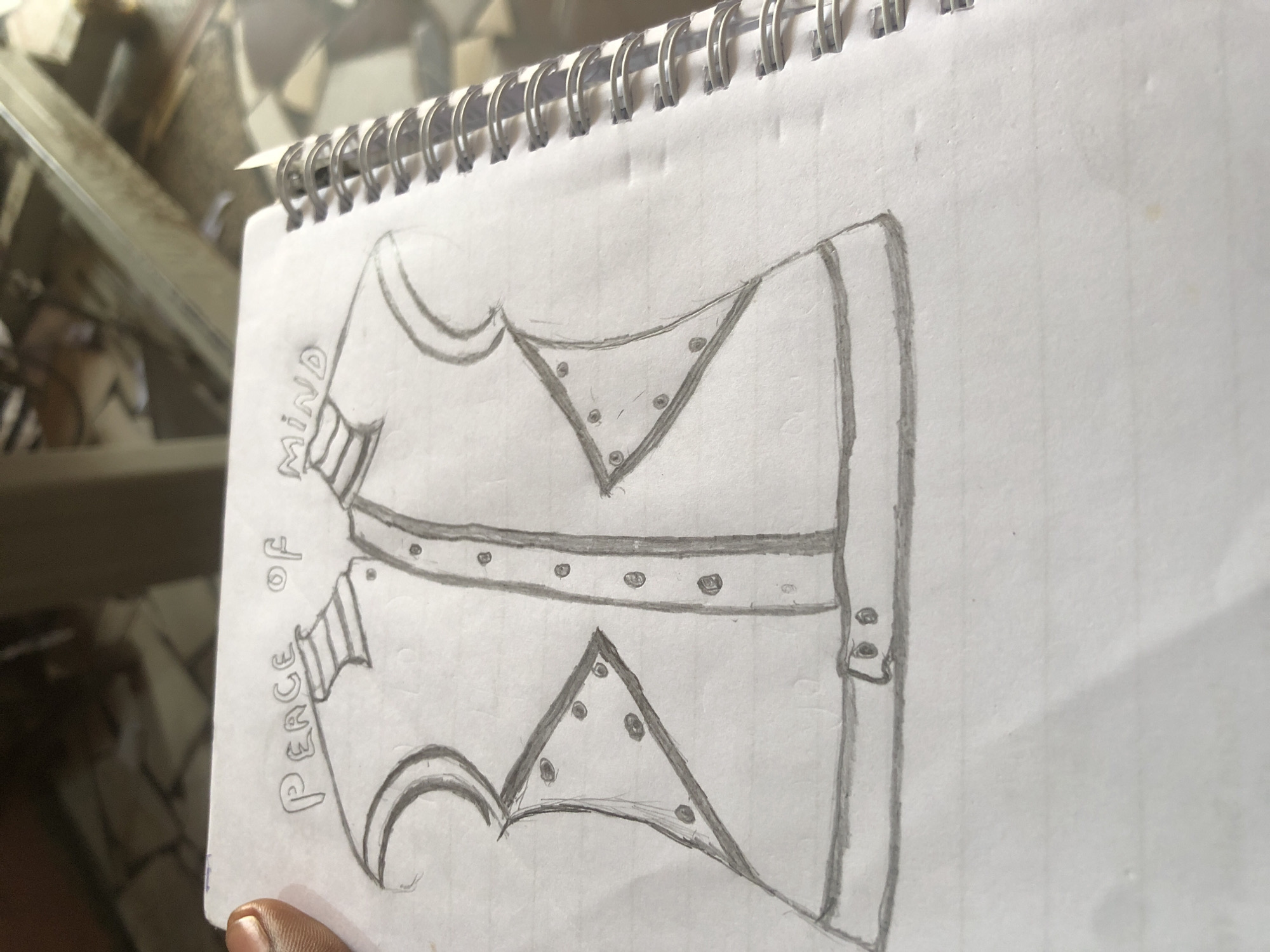 A pencil sketch of a vest is drawn on lined paper. The vest has a collar and buttons down the front. There are triangular decorative elements on the chest of the vest, each with several small circles within. A belt with a buckle is drawn around the waist of the vest. Text reading "PEACE OF MIND" is written above the vest. The paper is part of a spiral-bound notebook and appears to be on a table. The background is blurred and shows what appears to be a metal structure and possibly other objects. The scene is not located in Ado, Nigeria.
