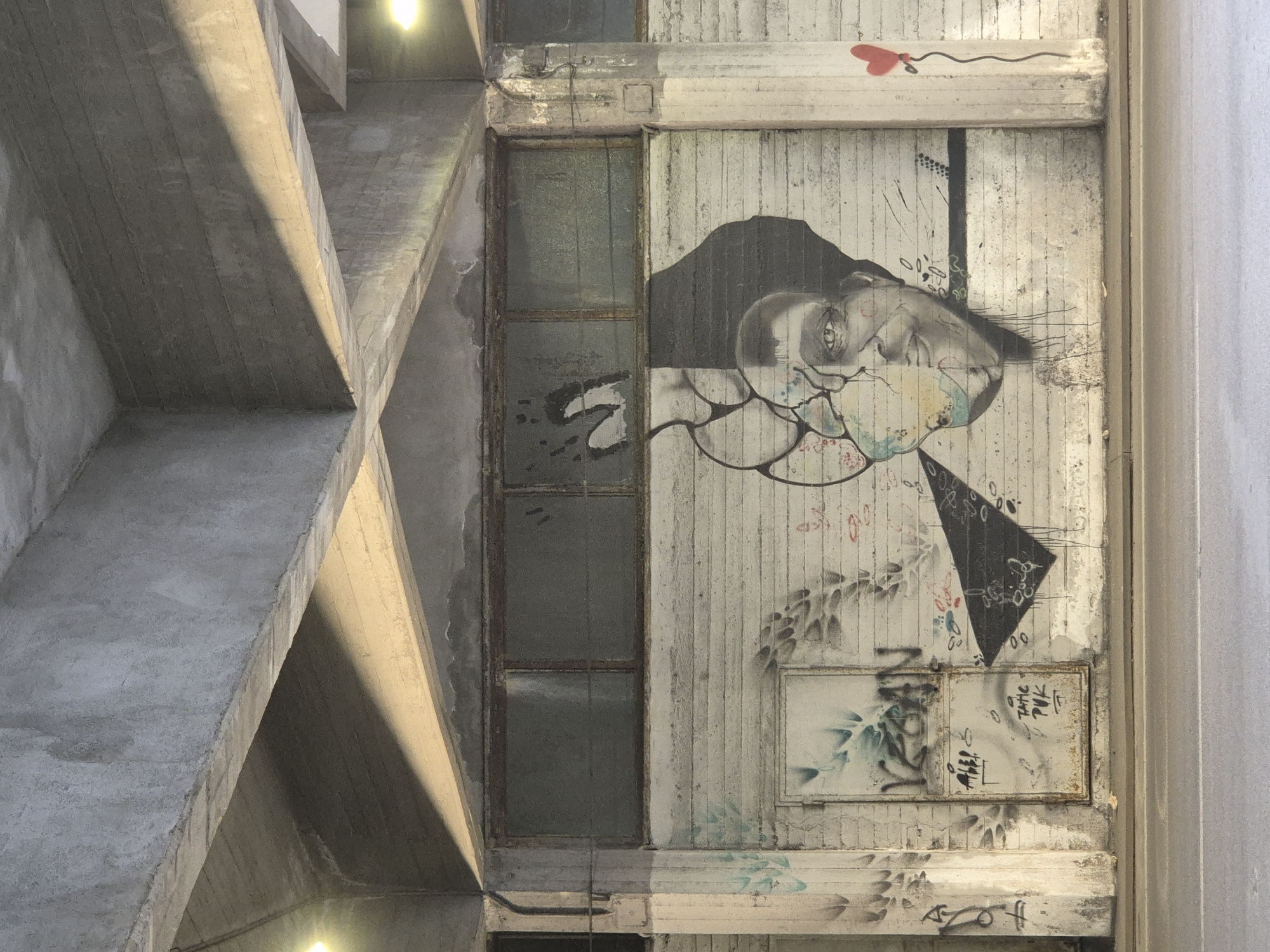 The image shows a section of a concrete structure, likely an underpass or bridge, in Taranto, Italy. The graffiti-covered wall features a large mural of a person's face, split down the middle. The left side of the face is fragmented and abstract, with colorful splashes and cracks. The right side is rendered in grayscale, with detailed features and a visible eye. To the left of the mural, there is a metal door with further graffiti. To the right of the mural, a simple drawing of a red heart with a string is visible. The concrete ceiling above the wall is textured and shows the marks of formwork. Harsh lighting creates strong shadows across the concrete surfaces, highlighting the textures and the graffiti. The scene is bordered by a road at the bottom of the frame.