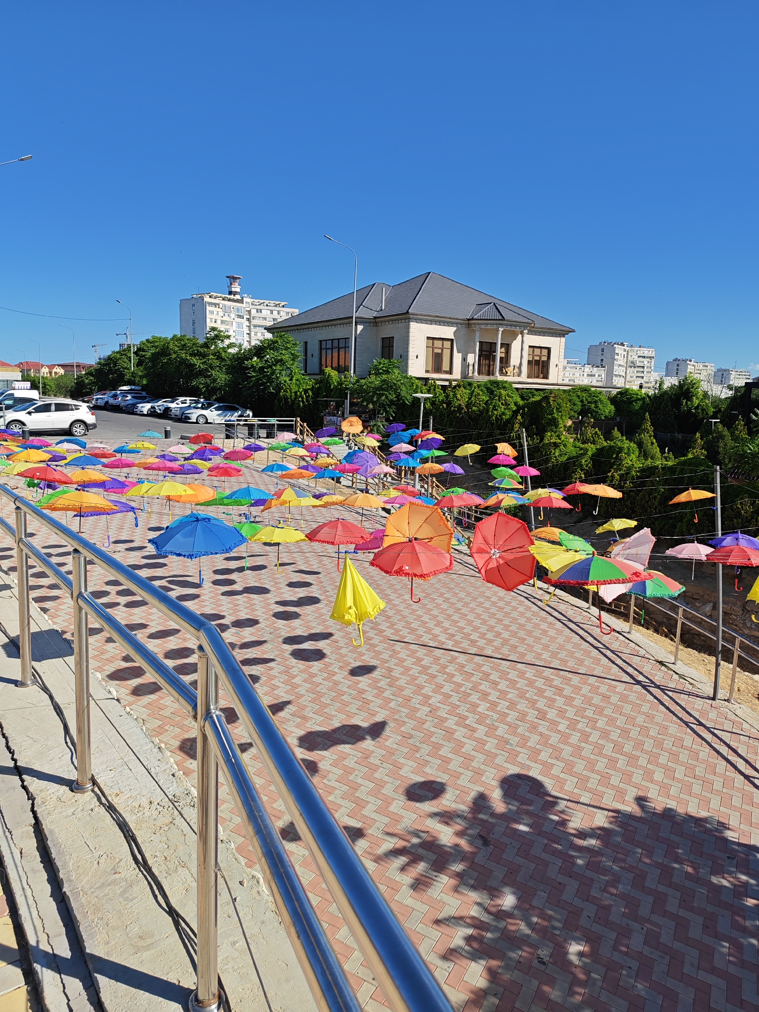 The image depicts an outdoor scene with a vibrant display of numerous colorful umbrellas suspended in the air, creating a visually striking installation. The umbrellas are of various colors, including red, orange, yellow, green, blue, purple, and pink, and are arranged at different heights, adding depth and dynamism to the scene. They are strung from wires or poles, forming a canopy over a pedestrian walkway paved with red and beige brickwork laid in a herringbone pattern.
In the background, a large, modern two-story house with a dark tiled roof and large windows is visible, situated on a slightly elevated area surrounded by lush green trees and bushes. Beyond the house, several multi-story apartment buildings are visible, indicating an urban or suburban setting. The sky is a clear, bright blue, suggesting a sunny day.
In the foreground, a silver metal railing runs along the left side of the image, bordering a concrete structure with visible cracks. The sunlight casts distinct shadows of the umbrellas and trees onto the paved ground, creating an interesting interplay of light and shadow.
To the left, several cars are parked on a paved surface, including a white SUV and other vehicles, suggesting proximity to a parking area.
The overall impression is one of an artistic public installation designed to add color and visual interest to a public space, likely a park, promenade, or a pedestrian zone in a city.
Based on external information, this installation of colorful umbrellas is located in the city of Aktau, Kazakhstan.