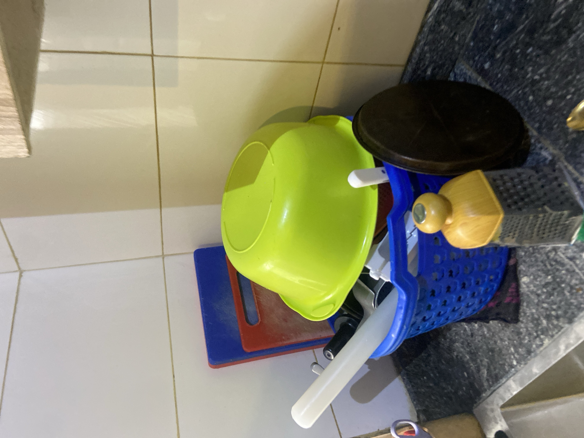 This image, captured in Howayi, Nigeria, presents a detailed view of a kitchen counter and adjacent wall. The background features a white tiled wall with light brown grout lines, extending upwards from the countertop. A wooden shelf, light in color, is partially visible at the top right of the frame. Against the tiled wall, two plastic cutting boards are propped upright; the front one is blue, and a red one is behind it. In the center, a bright lime-green plastic bowl is inverted and rests atop a blue, perforated plastic dish drying basket. The basket, positioned on the countertop, contains various kitchen utensils, including a prominent white rolling pin, several dark-handled knives or tools, and other white plastic utensils. The countertop is a dark grey surface with light speckles, suggesting a granite or similar material. Towards the bottom left, a portion of a stainless steel sink basin is visible. On the countertop in the foreground, a yellow and grey cheese grater with a wooden top stands vertically. To its right, a dark brown, round, flat object, possibly a pot lid or a small cutting board, is also positioned upright. A glimpse of a golden metallic object is visible in the bottom right corner. No individuals are present in the scene, and no actions are explicitly depicted.