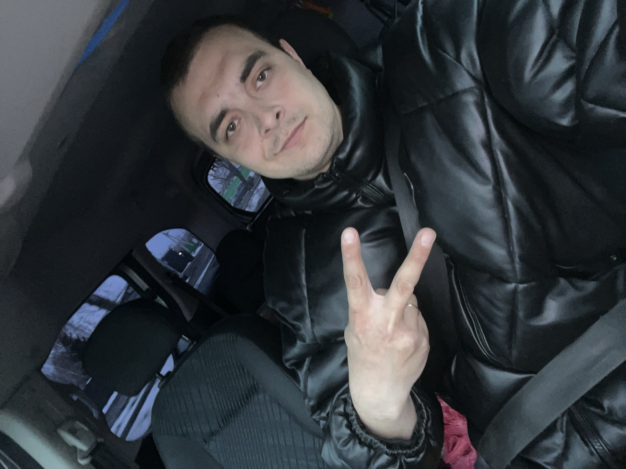 The image is a selfie of a man seated inside a car, likely in the front passenger seat. He has short dark hair, prominent dark eyebrows, and light brown eyes, with light stubble on his face. He wears a black, shiny puffer jacket and has a calm, slightly smiling expression while looking directly at the camera. His right hand is raised, forming a peace sign (V-sign) with two fingers, and a simple ring is visible on his right ring finger.
The interior of the car is visible, including a dark seatbelt across his chest and the textured fabric of the car seat to his left. Through the car windows, the exterior appears dimly lit, suggesting dusk, night, or a heavily overcast day. Patches of snow or frost are visible on the ground outside, indicating cold weather conditions. The overall mood conveyed by the man's expression and gesture is relaxed and positive. No visible text is present in the image. The location context provided is Novosinkovo, Russia, which aligns with the observed cold-weather environment.