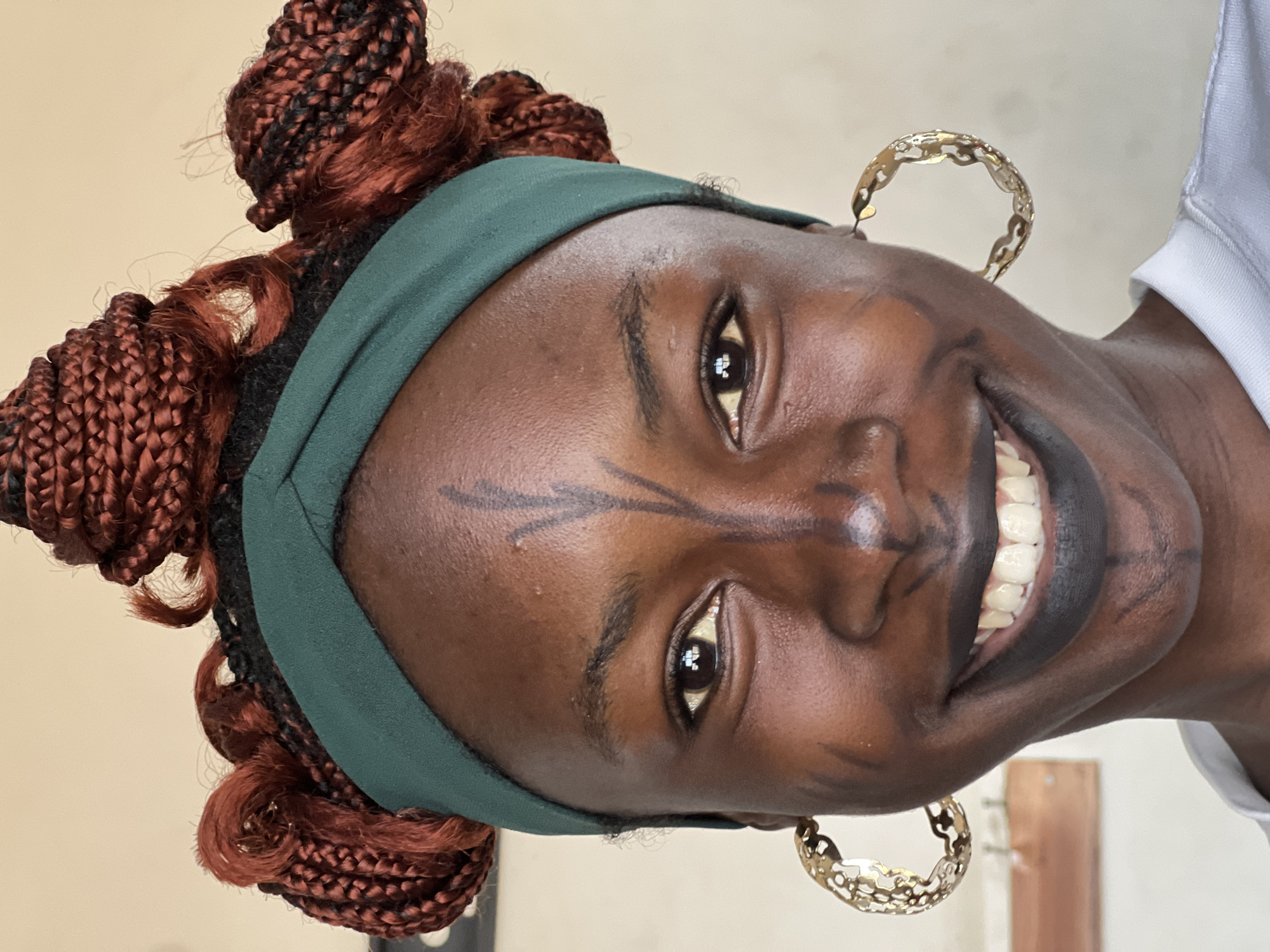 The image is a close-up, head-and-shoulders portrait of a young Black woman, likely taken outdoors in the city of Jos, Nigeria. She is facing forward and smiling directly at the camera. Her skin tone is a deep brown. She has her hair styled in numerous reddish-brown braids, some of which are intricately arranged in buns and curls around her head. A dark green headband is worn across her forehead, partially covering her hairline.
Her face is adorned with black face paint. A prominent vertical line extends from her forehead down to the bridge of her nose, with two smaller lines branching off from the top of the vertical line. A dot of paint is visible on her nose. Additional painted lines extend from the sides of her nose towards her cheeks, and also beneath her lips. She is wearing bold, matte black lipstick.
She has large, hoop earrings in her ears. The earrings are gold-colored and have an open, decorative design. Her eyes are open and dark, with a subtle smile playing on her lips, revealing white teeth. She is wearing a white top, with the collar visible at the bottom of the frame. The background is a plain, light-colored wall, with a hint of what appears to be a wooden shelf or surface on the left side, suggesting an indoor setting with natural light. The overall impression is one of cultural expression and personal style.
