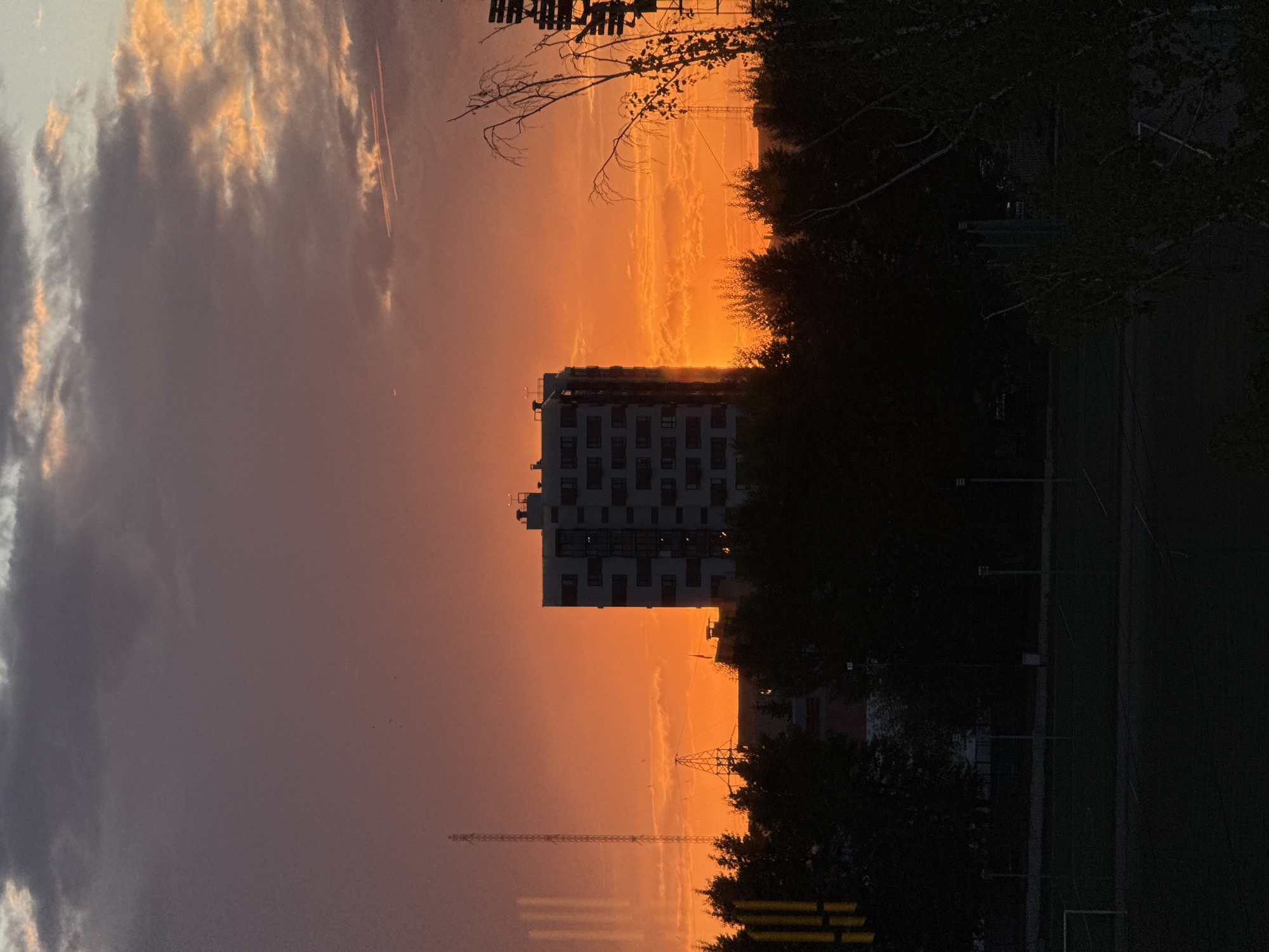 The image captures a sunset view from Krasnoyarsk, Russia. In the foreground, there's a tall building with multiple windows, situated between darker silhouettes of trees. The setting sun casts an orange glow behind the building, emphasizing the sky and cloud formations. Below, there is a tall cellular tower. A sports field is visible on the bottom right. The sky transitions from a dark gray-purple to an orange hue, with the brighter orange concentrated near the horizon where the sun is setting.