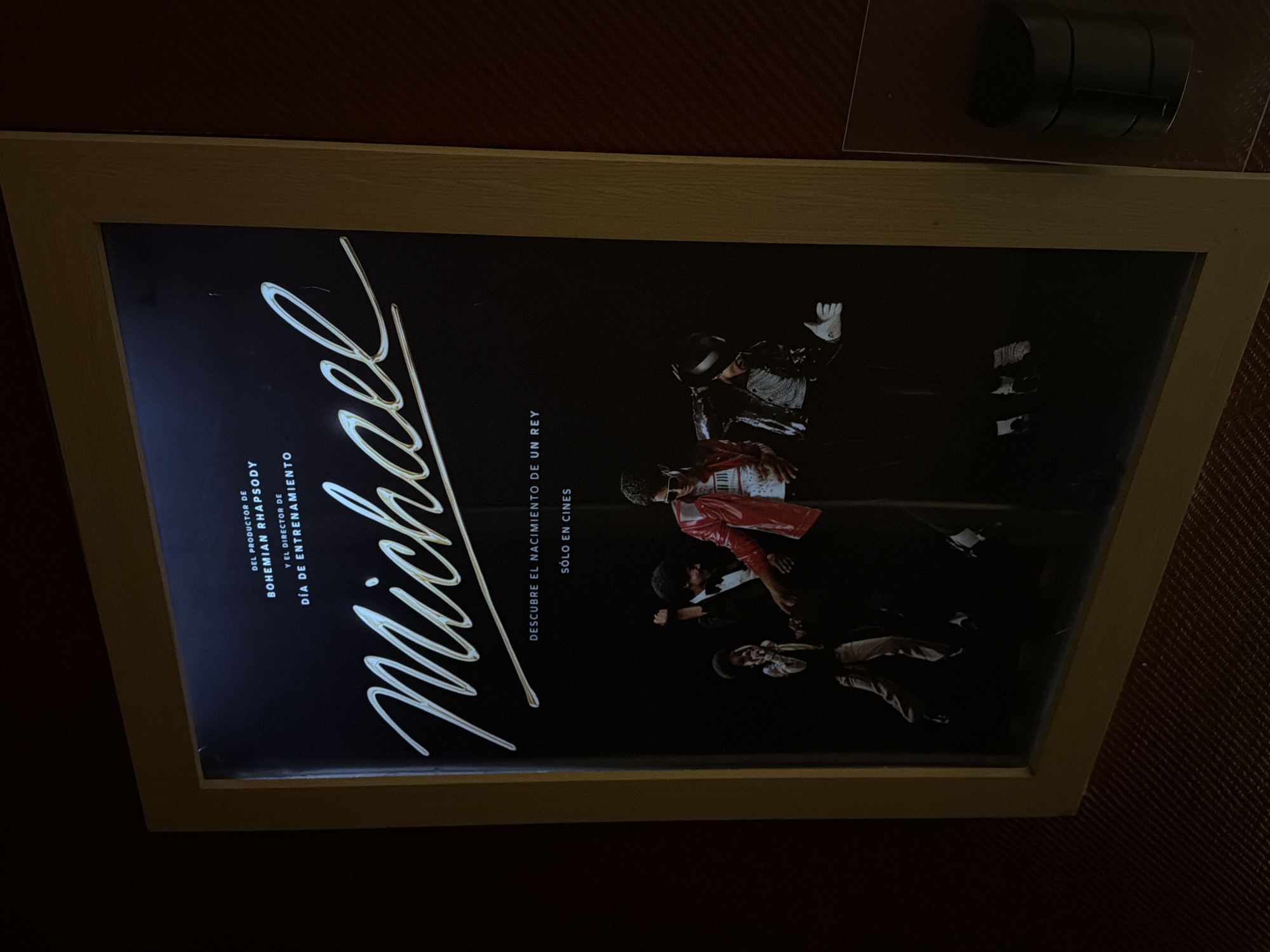 A framed movie poster for "Michael" is displayed against a red textured background. The poster features the name "Michael" in a stylized script font. Below the name are the phrases "DEL PRODUCTOR DE BOHEMIAN RHAPSODY" and "Y EL DIRECTOR DE DIA DE ENTRENAMIENTO". To the right, the text "DESCUBRE EL NACIMIENTO DE UN REY" and "SÓLO EN CINES" are visible. Images of Michael Jackson in different outfits are displayed on the poster. A black cylindrical object is positioned above the framed poster. The location is identified as Unknown, Unknown.