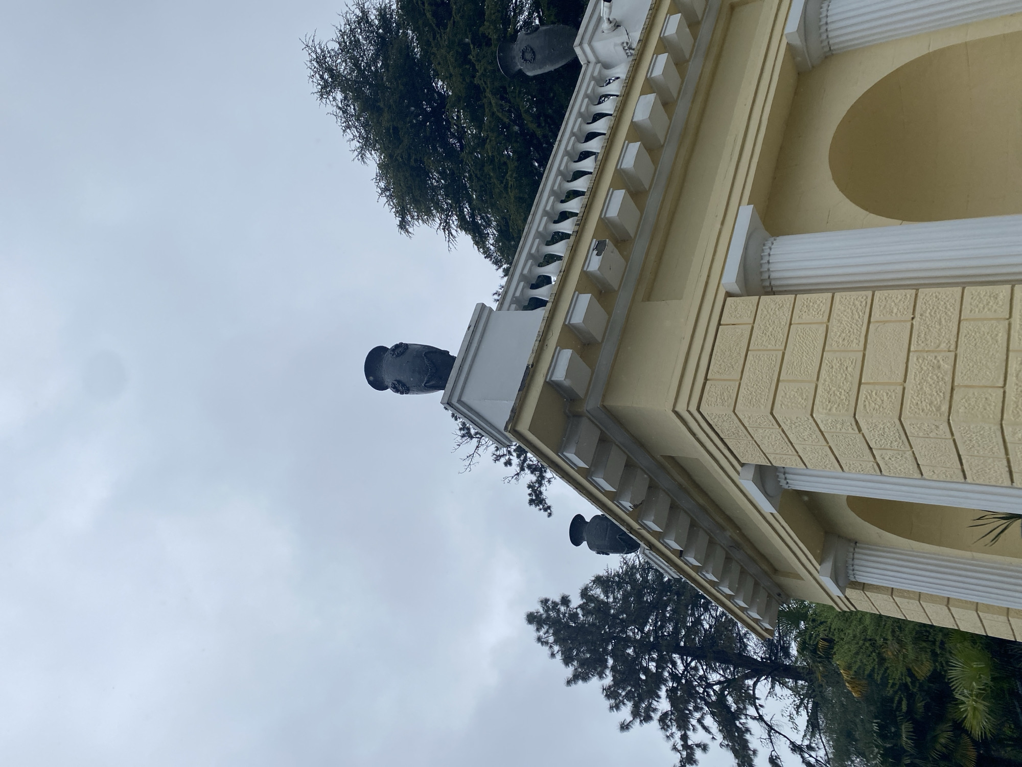 A low-angle shot depicts a section of a building in Sochi, Russia. The building is characterized by yellow walls and white architectural details, including fluted columns, decorative trim, and statues atop a balcony. The statues appear dark against the light sky. The sky is overcast, with shades of gray. In the background, trees with dark green foliage are visible. The perspective of the shot creates a sense of looking up at the building and the sky. The overall composition is one of architectural detail against a natural backdrop.