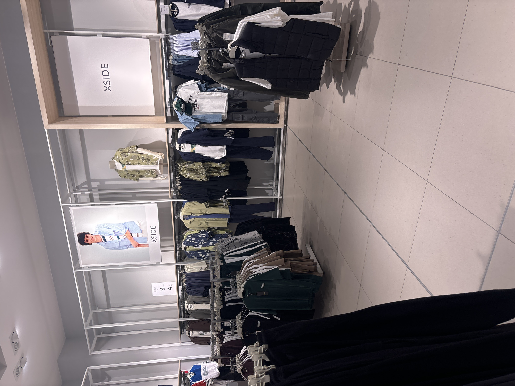 The image shows a clothing rack inside a store, presumably in the city of Unknown, Unknown. The rack is organized with shelves displaying folded and hanging clothing. Signage on the rack features the brand name "XSIDE". The clothing includes a variety of styles, from casual shirts and pants to jackets and vests. The rack is constructed of metal and has a light wood frame. The floor of the store is tiled. The image is rotated, causing the items to appear sideways.