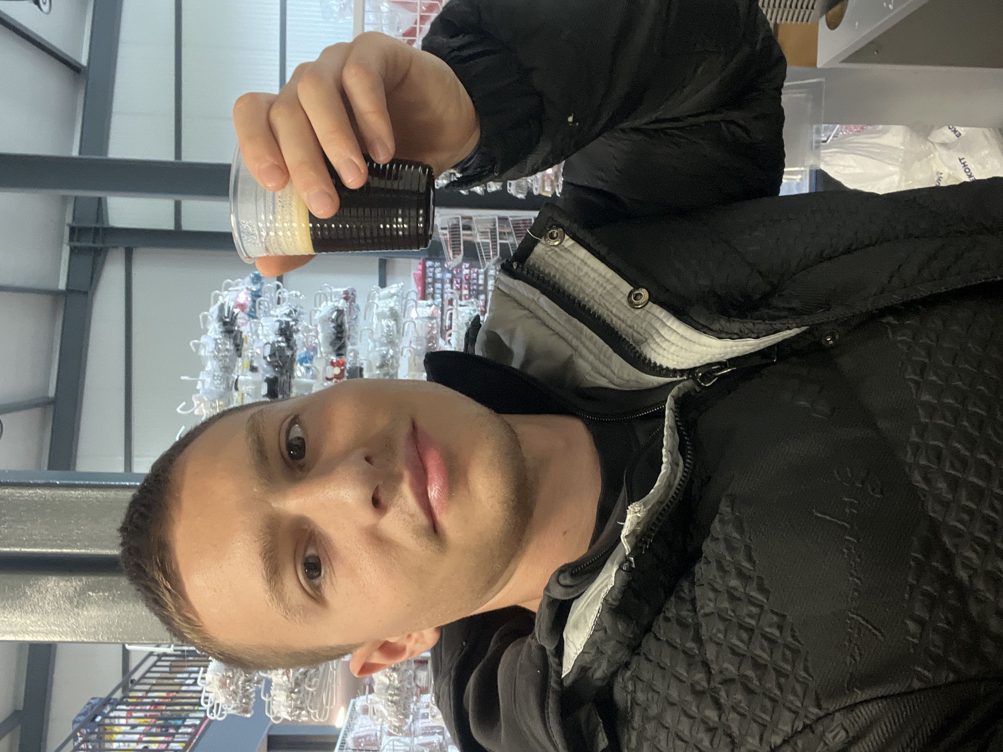 A man stands indoors, holding a clear plastic cup with dark liquid inside. The cup appears to contain coffee. The background features rows of items displayed on shelves. The exact location in Burgas, Bulgaria cannot be determined from the image.