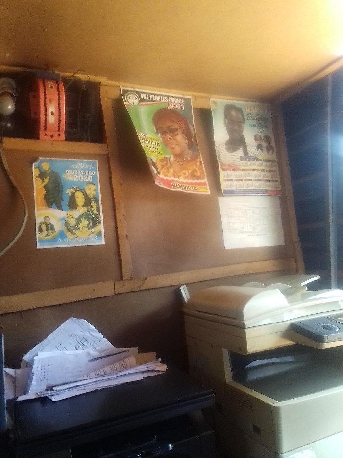 The image shows a cluttered office space in Auchi, Nigeria (latitude: 7.063, longitude: 6.274). Three posters are mounted on the wall: "The Peoples Choice 2021" featuring a woman in a headscarf, a poster with the words "Happy Birthday" and a poster advertising a concert by "Chizzy Ugo" in 2020. A large stack of paper is piled on a black office desk next to a multifunction printer/copier. The office appears to be in operation. There is a ceiling light and electrical wires visible, indicating a working office environment. The interior wall is painted a light yellow and the wall is made of wood. The room appears to be dimly lit, possibly in the afternoon.