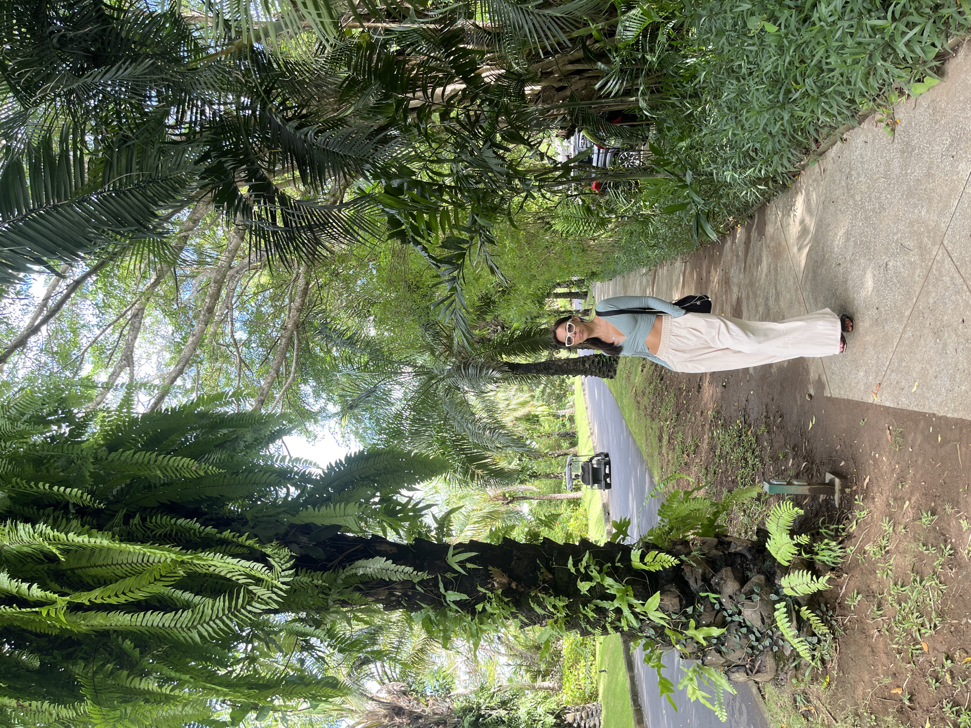 A person stands on a gray sidewalk in a lush, tropical location. They are wearing a light blue top, white pants, and white sunglasses. A black bag hangs on their shoulder. The sidewalk is bordered by dense greenery including palm trees and ferns. A paved road runs parallel to the sidewalk, and a dark-colored golf cart is visible on the road. The scene evokes a tropical resort setting. The location is in Kuta Selatan, Indonesia.