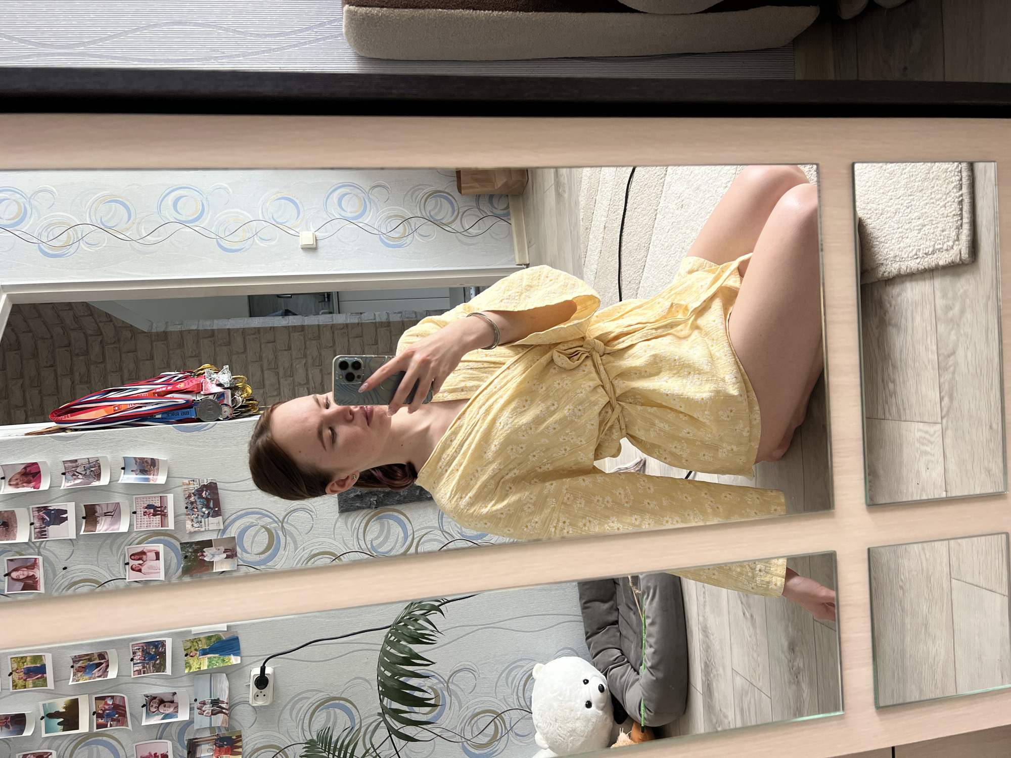 A young woman is seen in a mirror selfie. She is wearing a yellow, floral-patterned robe and holding a smartphone in her right hand. The mirror reflects a room with a wall adorned with photographs, an electrical outlet, and what appears to be a houseplant. Also visible are some medals and ribbons on a shelf. In the reflection are a stuffed animal, a pet bed, and hardwood flooring. The location is Chaikovsky, Russia.