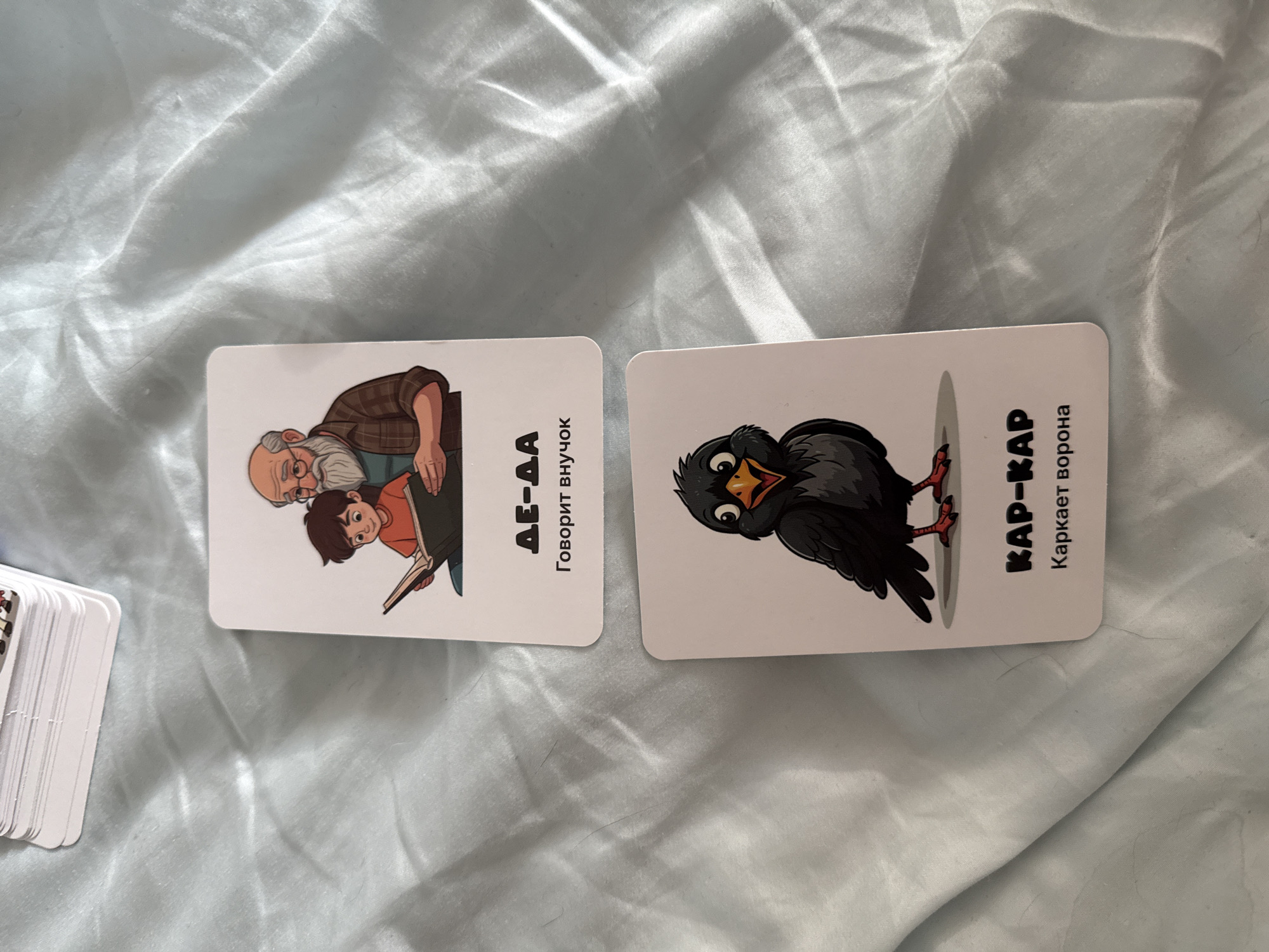 The image shows two cards lying on a white fabric.
The card on the left features a cartoon illustration of a grandfather and his grandson reading a book. The card also has the text "ДЕ-ДА Говорит внучок".
The card on the right shows a cartoon illustration of a crow. The card also has the text "КАР-КАР Каркает ворона".
The cards are placed on a rumpled white fabric. A stack of similar cards can be seen in the bottom left corner of the image.
