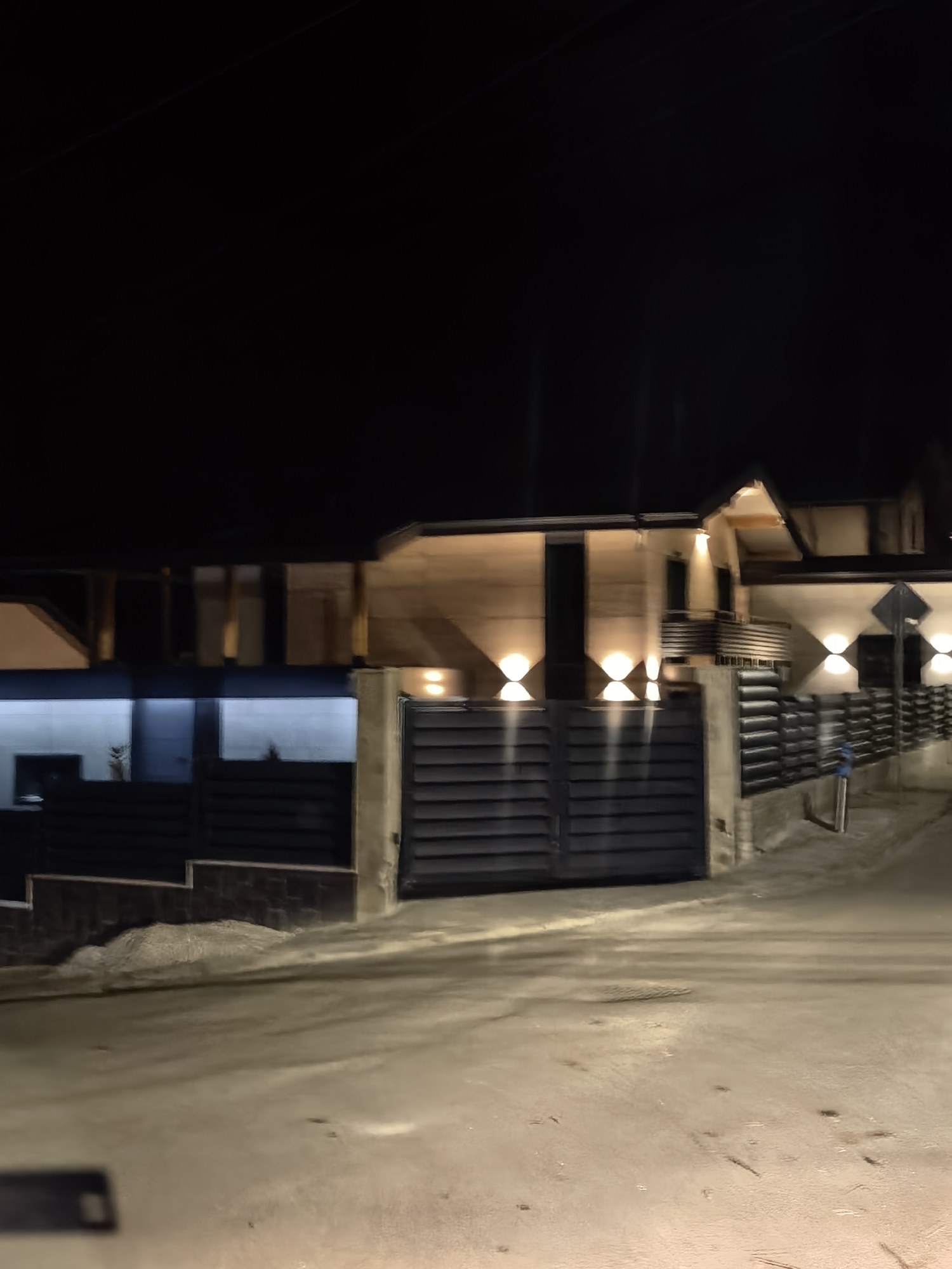 The image captures a nighttime scene of a modern house situated in an urban environment, likely residential. The house is characterized by a combination of horizontal and vertical lines, a flat roof section to the left, and a gabled roof section to the right. The exterior walls appear to be constructed of a light-colored material, possibly concrete or stucco. The house is lit by multiple exterior lights, some of which are placed symmetrically, creating a distinct illumination pattern on the walls and emphasizing the architectural features. A dark gate is visible in the front, and a low fence extends on either side, leading down to street level. The road in front of the house is paved and appears to be slightly wet or reflective. The background is dark, suggesting that the image was taken at night. There are no people visible in the image. The location is specified as Unknown, Unknown, which indicates that the exact location in the city is unknown.