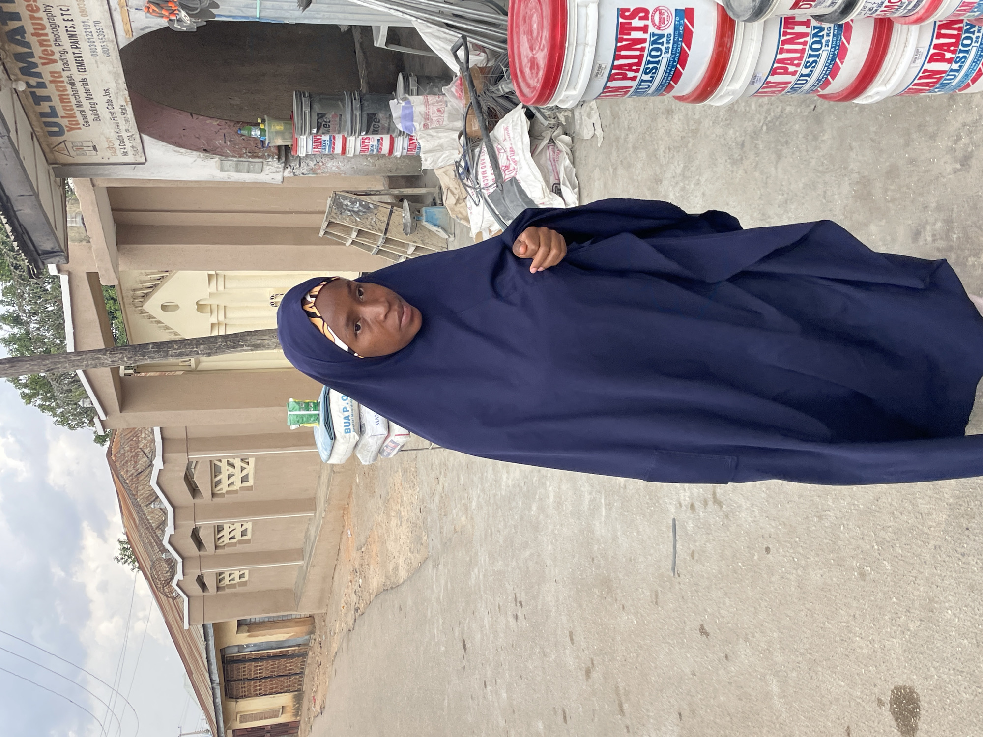 A woman in a dark blue hijab stands on a concrete surface outdoors. Behind her is a building with a light brown facade, notable geometric patterns, and a rusty corrugated metal roof. Paint buckets are visible behind her, with branding indicating the "DAN PAINTS" brand. A sign for "Yakamata Ventures" is partially visible, advertising general merchandise, trading, photography, and building materials. The sign indicates a location in Jos, Plateau State, Nigeria. Other building materials, including cement, and bags of what appear to be building supplies are in the background. The sky is overcast.