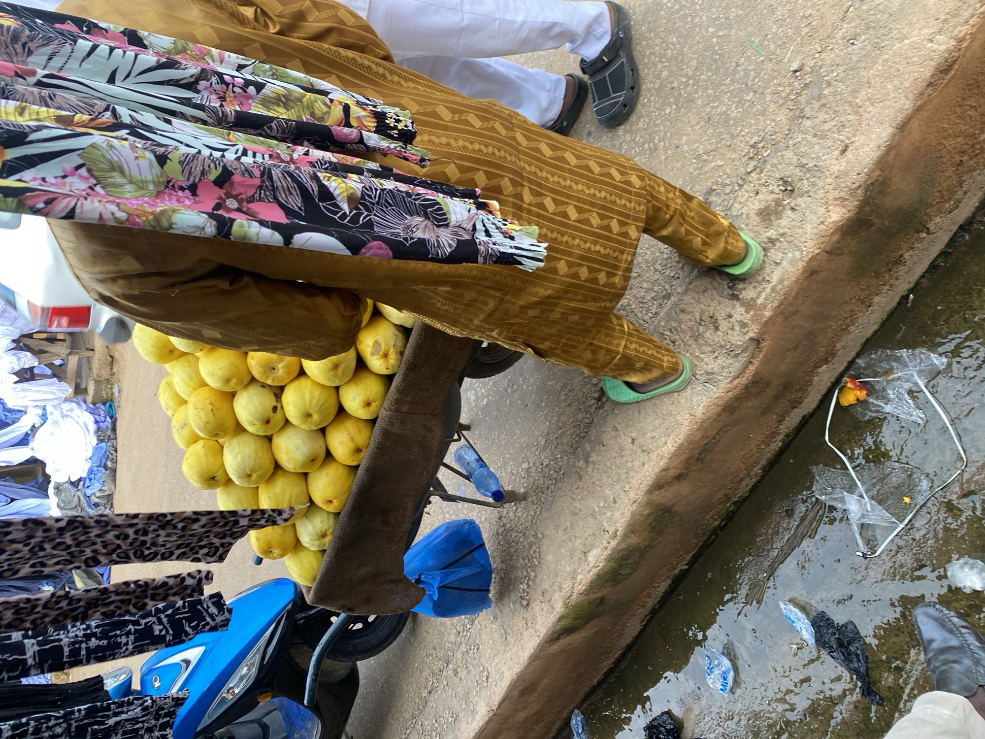 In a street scene in Jos, Nigeria, a street vendor is preparing to sell produce. A cart laden with yellow fruits, possibly guavas, is positioned beside a blue scooter. The vendor, adorned in a traditional brown patterned outfit and a colorful scarf, stands next to the cart. A person in white trousers and black crocs stands to the right. The ground is paved with concrete, and a small, dirty gutter runs along the side, filled with water and debris, including plastic bags and a discarded water bottle. In the background, laundry hangs on a line, suggesting a residential or commercial area. The overall scene depicts a bustling urban environment with elements of commerce and daily life.