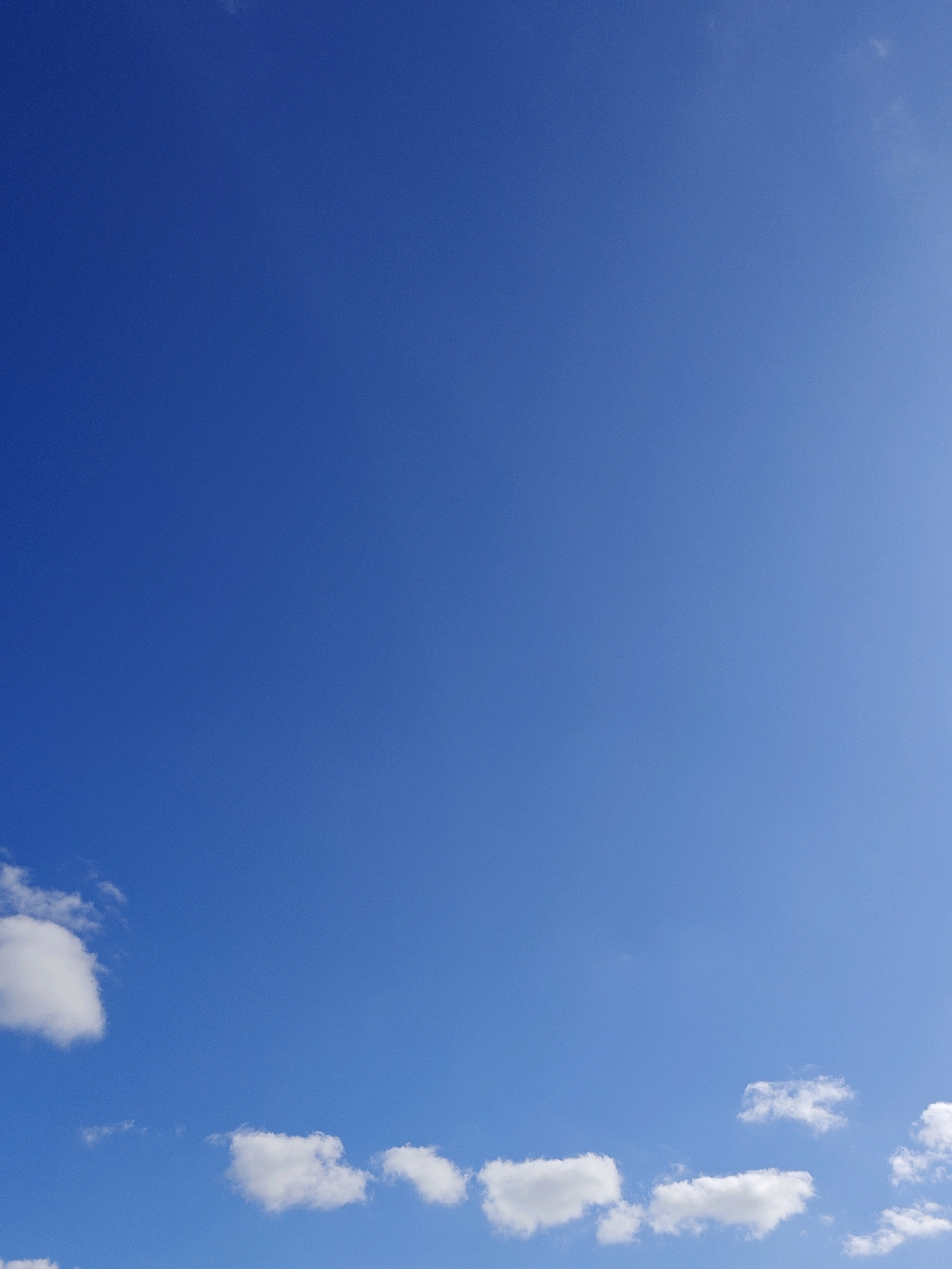 The image depicts a clear blue sky with scattered white clouds near the bottom edge of the frame. The sky occupies the vast majority of the image, with a gradient of blue that is lighter towards the right side and darker towards the left. Several cumulus clouds are visible, with varying degrees of density and shape. On the far left, a larger, fluffy cloud is prominently featured. To the right of this, smaller, more wispy clouds are scattered across the lower portion of the sky. The scene appears to be an outdoor view, consistent with a sunny day in Trubino, Russia, though no specific landmarks or human activity are visible.