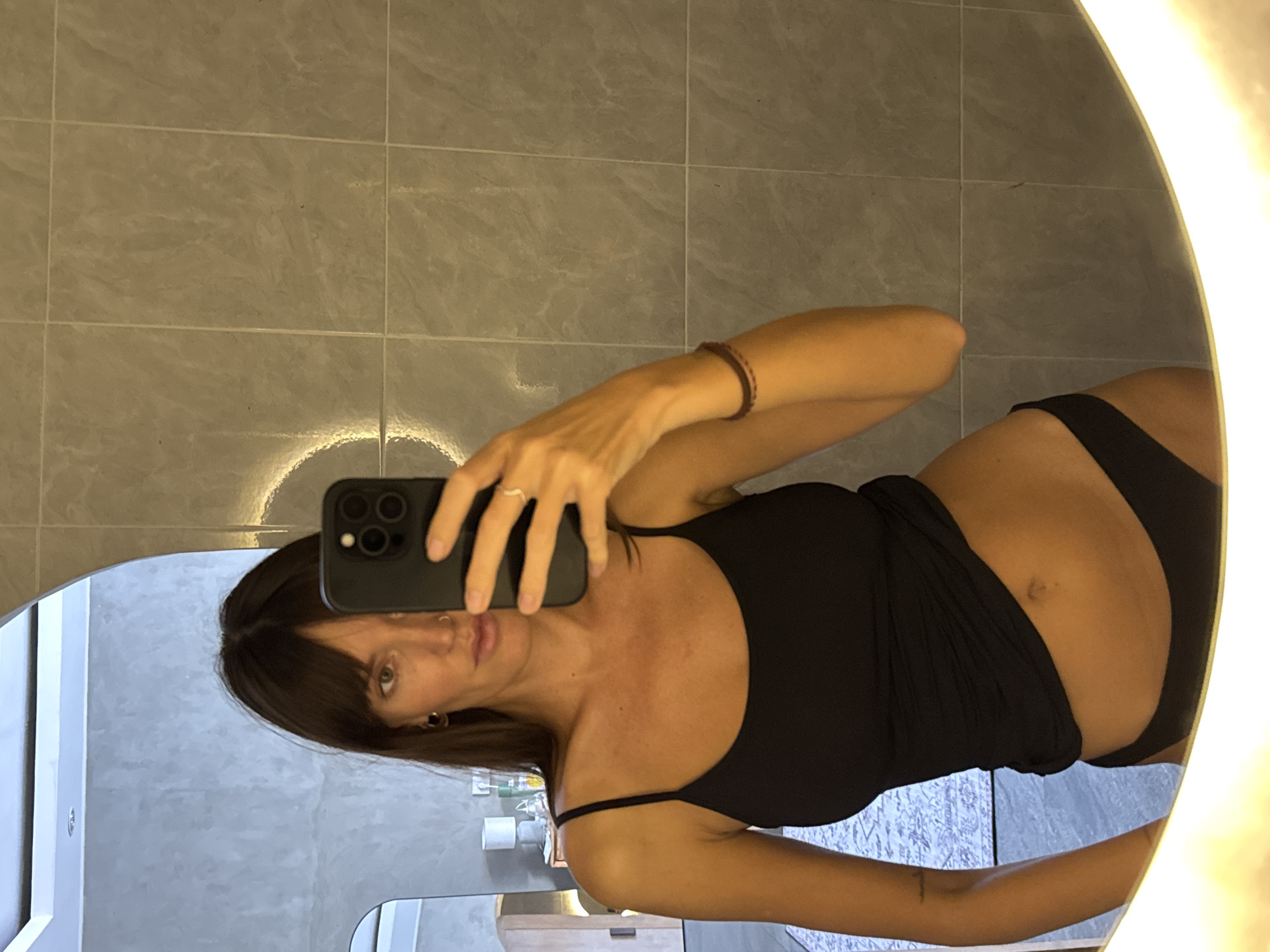 A woman with brown hair and bangs takes a selfie in a round mirror in what appears to be a bathroom. She holds a black smartphone in her right hand. She is wearing a black camisole and underwear. The bathroom has gray tiled walls and a white vanity. The location is suspected to be in North Kuta, Indonesia.