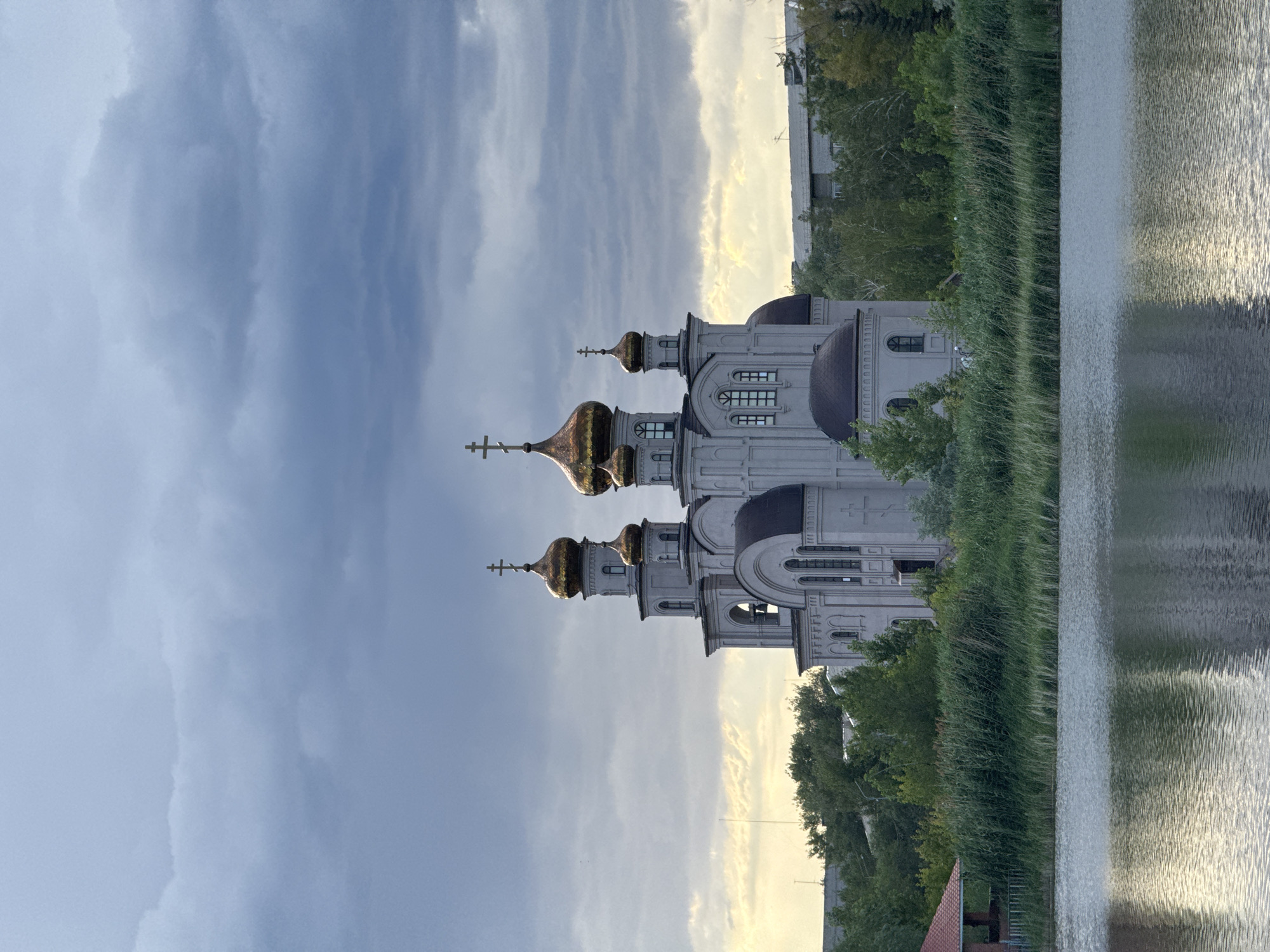 This media file depicts an Orthodox church situated beside a body of water, likely a river or lake, in Pavlodar, Kazakhstan. The church is a prominent structure with multiple domes, some of which are golden and reflect the light. It features traditional architectural elements such as arched windows and decorative molding. Surrounding the church are lush green trees and tall reeds growing along the water's edge. The water itself is calm, with ripples creating a textured surface that reflects the sky and surrounding greenery.
The sky is partly cloudy, with dramatic cloud formations suggesting either an approaching storm or the aftermath of one. The lighting indicates it is likely late afternoon or early evening, given the soft, diffused sunlight and the warm tones in the sky, particularly on the right side of the frame. There is no visible text in the image. The scene evokes a sense of tranquility and spiritual contemplation, enhanced by the natural beauty of the setting and the imposing presence of the church. The overall impression is one of a peaceful, perhaps slightly somber, moment in time.