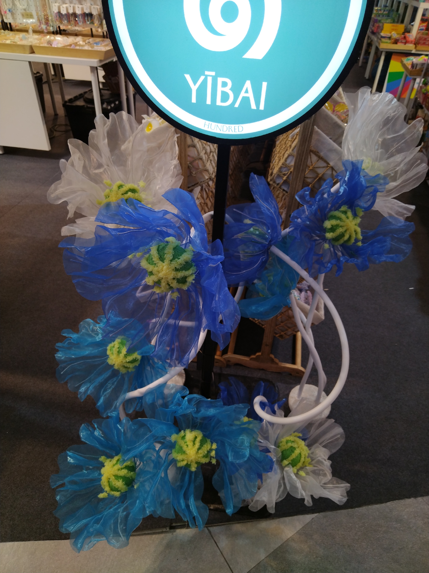 The image shows a display of artificial flowers in blue, white, and yellow, with a turquoise circular sign that reads "YIBAL HUNDRED" in white text. The flowers appear to be made of translucent plastic or fabric and are arranged on a white structure. In the background, there are shelves with various items, possibly in a retail setting. The floor is a dark gray carpet. This scene is likely from a shopping area or market in Patong, Thailand.
