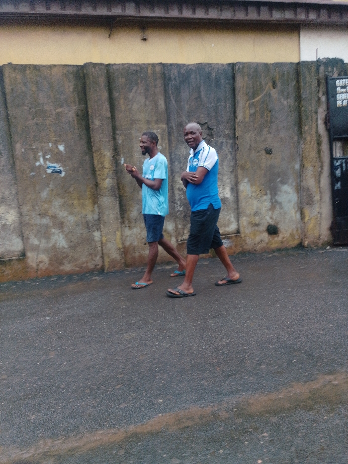 The scene depicts two adult males walking side-by-side on a damp asphalt road in the city of Agani, Nigeria. Behind them is a weathered, segmented concrete wall, topped by a section of a yellow-painted building. To the far right, a dark gate with an illegible white sign marked "GATE" is partially visible.
The male on the left, with a short beard, wears a light blue short-sleeved t-shirt, dark shorts, and light blue flip-flops. His hands are raised in front of him, palms slightly open, and he is smiling, looking to his right. The male on the right, bald, wears a blue and white collared polo shirt with a crest on the left chest, black shorts, and black flip-flops. His arms are crossed over his chest, and he is smiling while looking directly towards the observer. Both individuals are in mid-stride, moving from left to right across the frame.