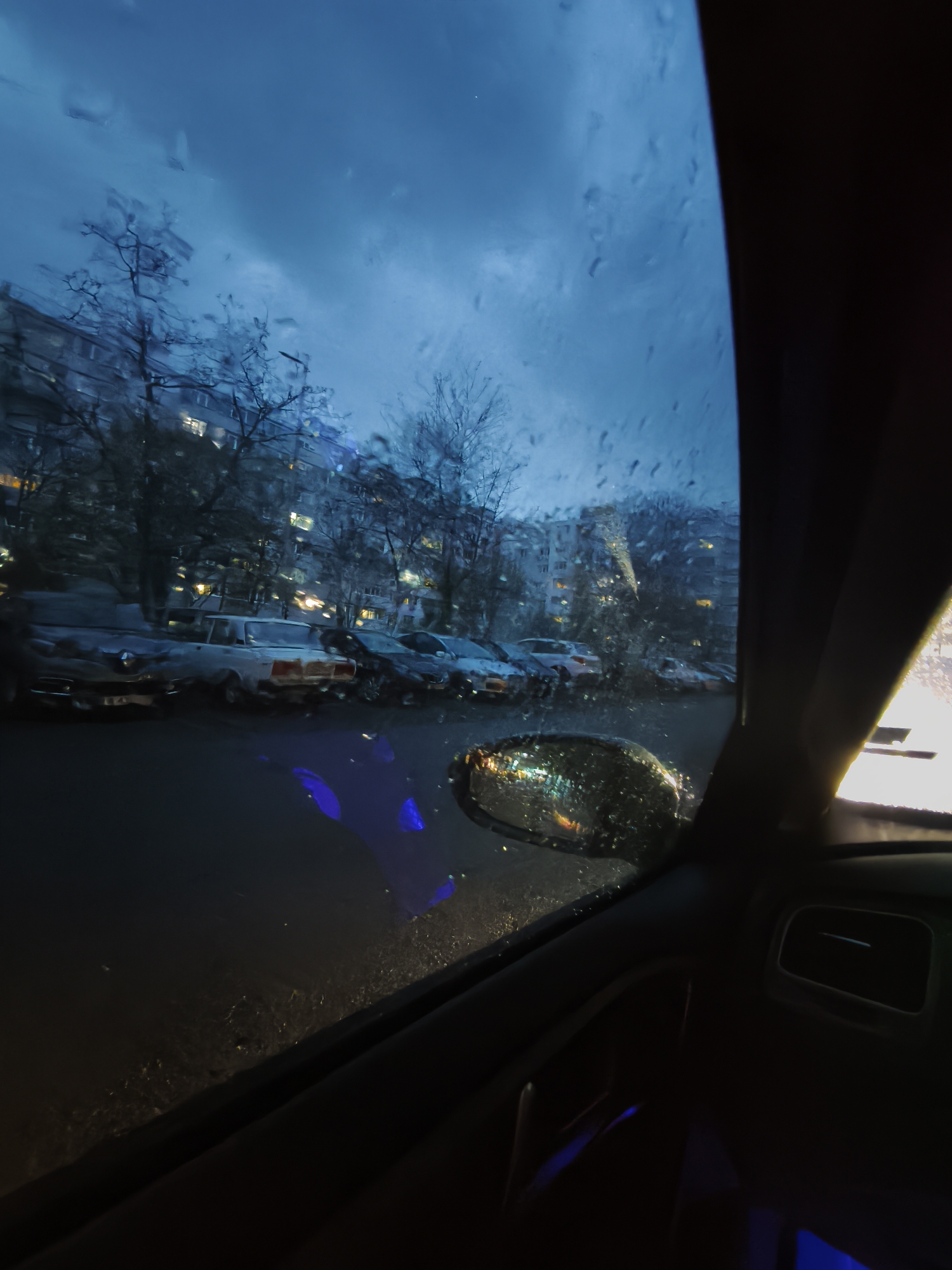 This vertical image depicts a street scene in Burgas, Bulgaria, viewed from inside a car through a rain-splattered windshield. The time of day is night or very late dusk, characterized by a dark blue, cloudy sky and illuminated windows in background buildings.
In the immediate foreground, to the right, a significant portion of the car's interior is visible, including the dark A-pillar, part of the dashboard, and a side mirror heavily coated in raindrops, reflecting distorted street lights. Hints of blue ambient lighting are present inside the car, subtly illuminating parts of the interior and reflecting on the lower section of the windshield.
The midground reveals a residential street lined with numerous parked cars. A prominent older model white sedan, possibly a Lada or similar classic vehicle, is clearly visible among others. Bare-branched trees stand along the roadside, their silhouettes adding to the somber mood. Beyond the street, multi-story apartment buildings rise into the frame, their windows emitting warm yellow light, contrasting with the cool blue tones of the exterior scene.
The entire outside view is obscured and softened by countless raindrops on the windshield, indicating ongoing rain or recent precipitation. The scene conveys a quiet, perhaps reflective, moment from within a vehicle, observing a typical urban residential environment under rainy, nighttime conditions. No discernible text is visible within the image.