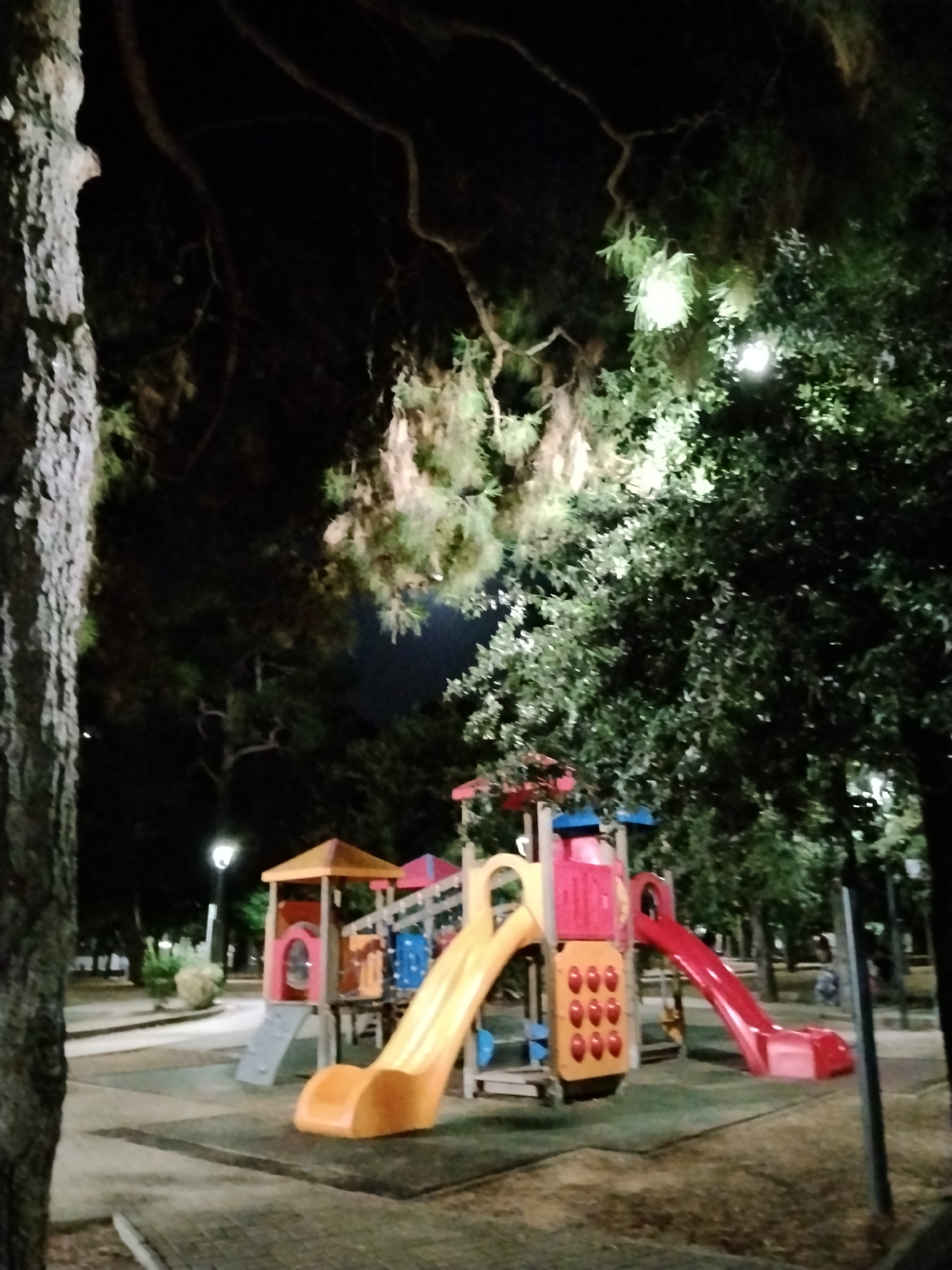 The image depicts a playground in Bitonto, Italy, at night. The playground features a red and yellow slide, a climbing structure with colorful elements, and a gray ramp. The ground beneath the playground equipment appears to be a dark rubberized surface. Surrounding the playground are trees with lush green foliage, illuminated by artificial lighting, creating a somewhat ethereal atmosphere. In the background, a lamppost with a light is visible, and a few indistinct figures can be seen walking in the distance. The overall impression is that of a tranquil, illuminated public space.