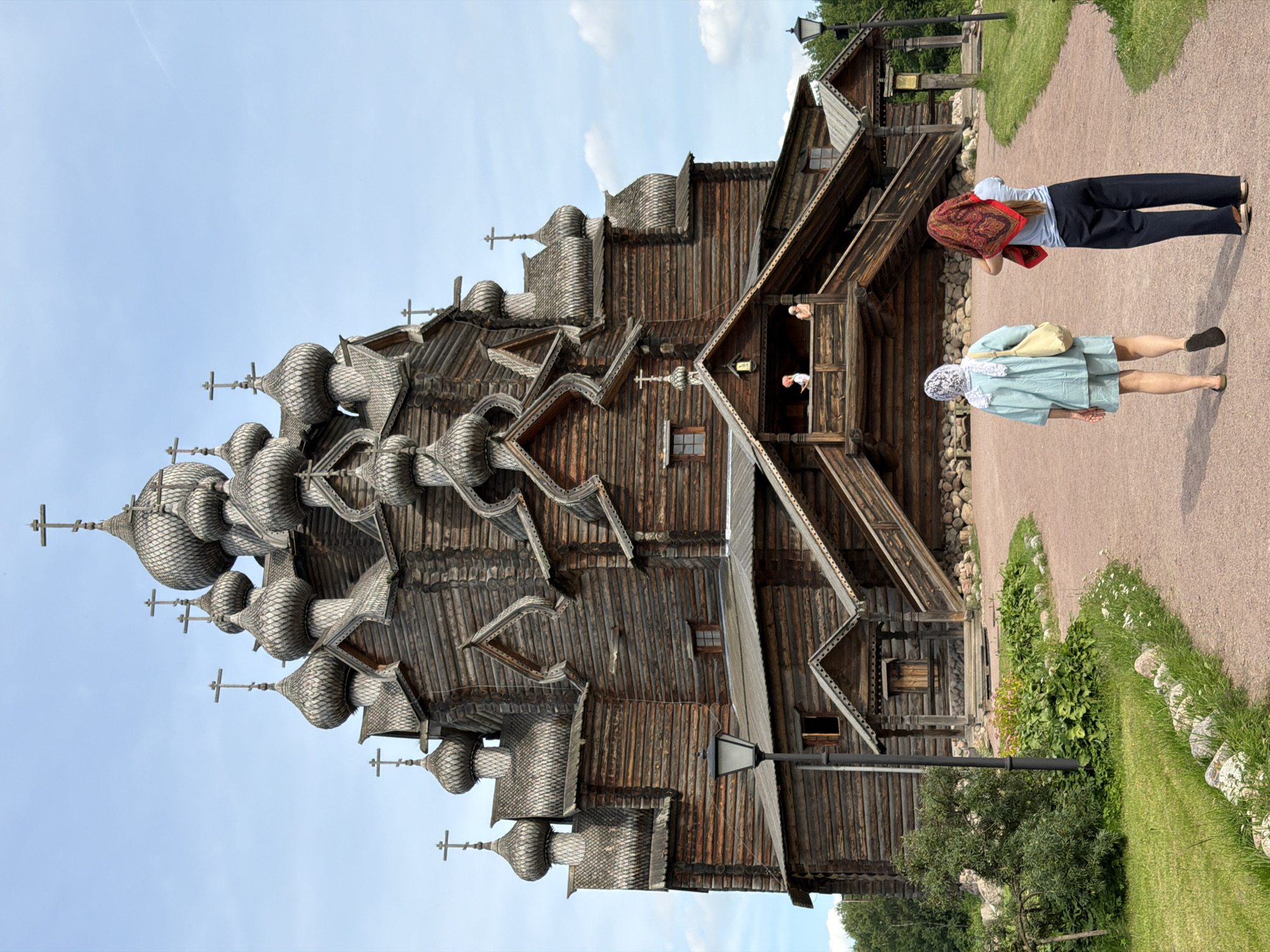 In the city of Imeni Sverdlova, Russia, a large, ornate wooden church stands against a bright blue sky with scattered white clouds. The church features multiple onion domes, each topped with a cross, and is constructed from dark, weathered logs. Decorative wooden carvings adorn the rooflines and eaves. A wide, gravel path leads towards the church, with grassy areas and some greenery on either side. Two women are walking away from the viewer on the path towards the church. The woman in the foreground on the left wears a light blue outfit and a white headscarf. The woman on the right wears a red and patterned scarf covering her head and a blue shirt with dark pants. Several people are visible on a wooden balcony on the church's facade. A traditional lamppost stands on the left side of the path.