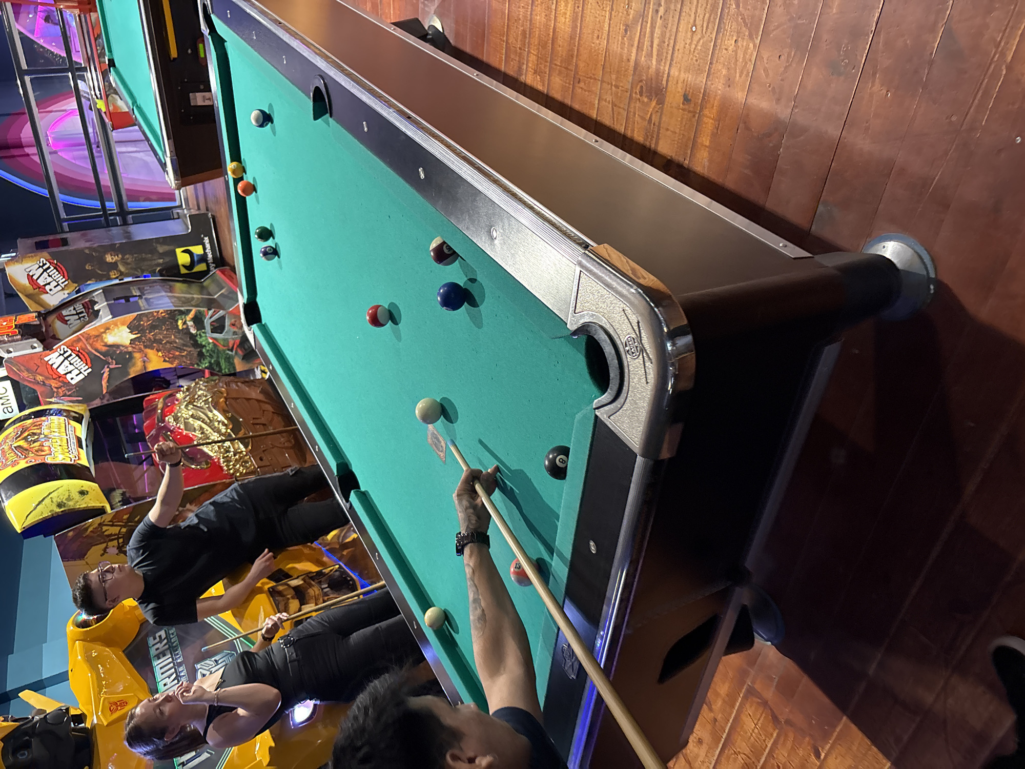 The image shows a billiards table in an entertainment establishment. The table is green and has several billiard balls on it. Two people are standing around the table, one of whom is holding a cue stick and appears to be aiming at a ball. Arcade machines are visible in the background. The specific location in Villa General Belgrano, Argentina, cannot be determined from the image alone.
