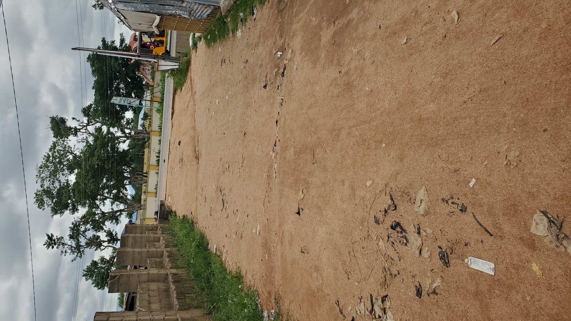 An outdoor, medium-range shot captures a dusty, unpaved road leading into a clearing. The road is sparsely littered with debris, including dark plastic bags and some dry leaves. To the left, a low, rough-hewn brick wall borders the road, with overgrown grass peeking out from its base. In the background, several large trees with full green canopies dominate the landscape, contrasting with a pale, cloudy sky. Beyond the trees, buildings with yellow trim are visible, suggesting a more developed area. A yellow auto-rickshaw is parked near the right edge of the frame, partially obscured by a metal barrier. A signpost stands near the auto-rickshaw, its text illegible at this distance. The overall impression is a rural or semi-urban environment with signs of both natural elements and human activity.
